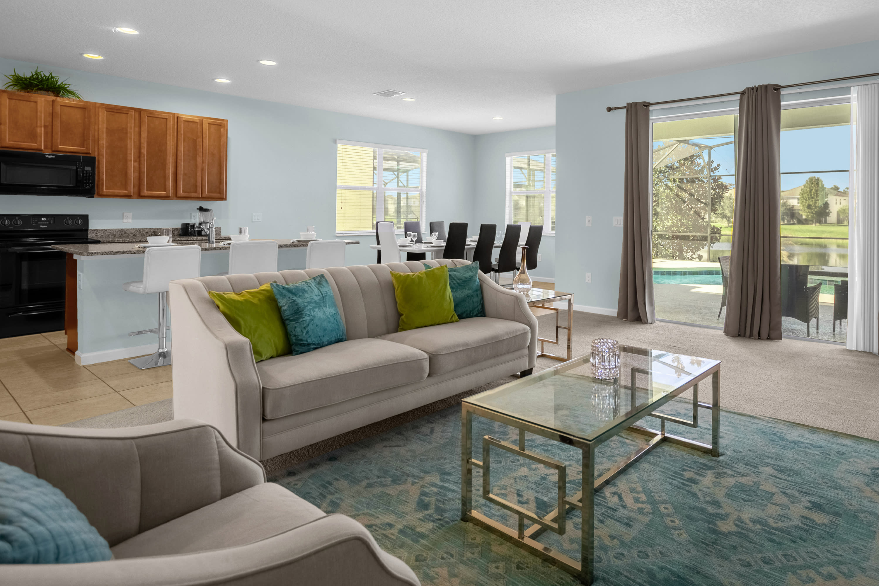 The open-concept living room features a beige sofa with colorful throw pillows and a glass coffee table on a blue patterned rug. Sliding glass doors lead to the outdoor pool area, and a dining area with a long table is visible in the background.