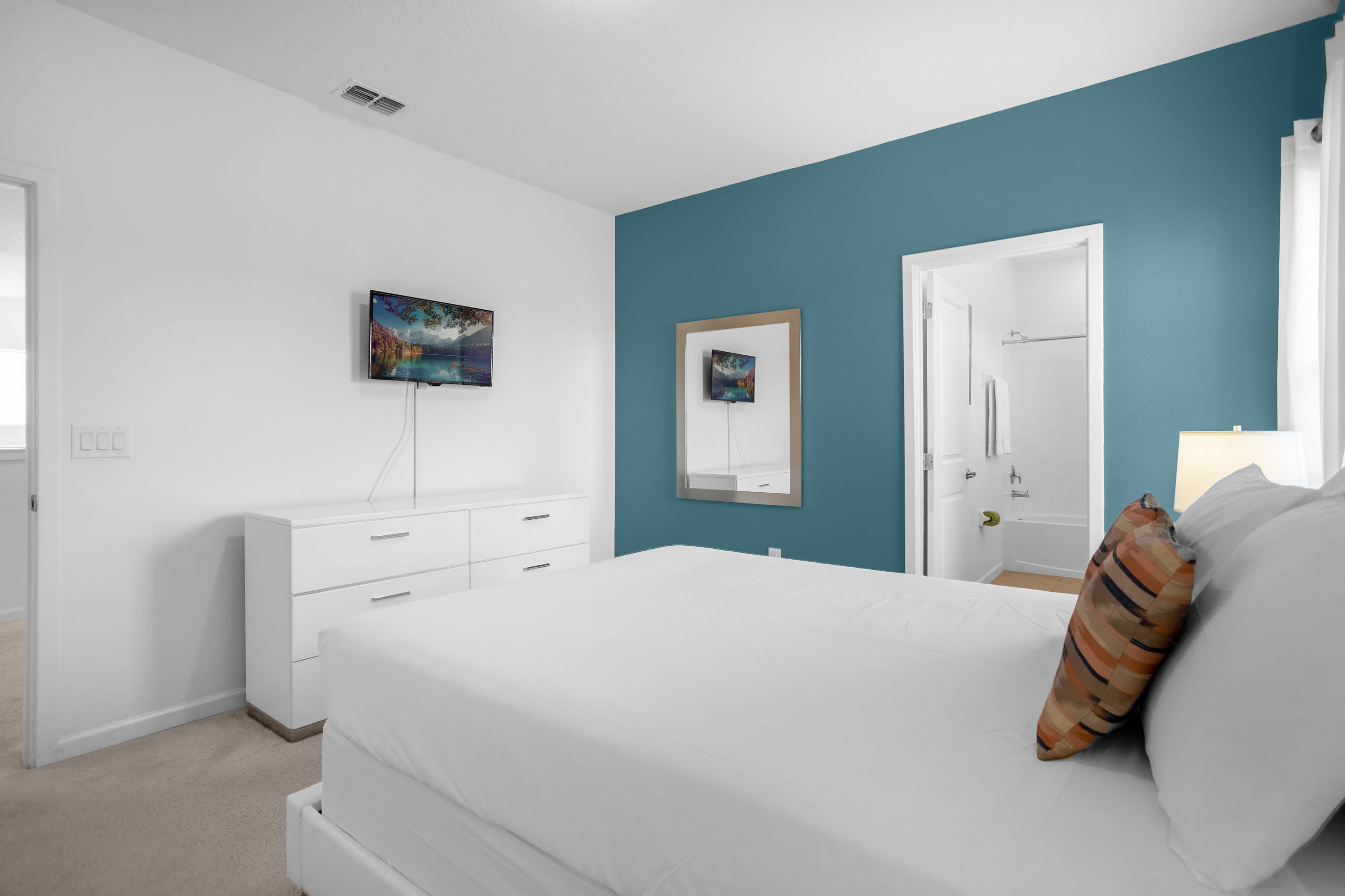 View of a bedroom with a white queen-sized bed, a white dresser with a wall-mounted TV, and a teal accent wall. A mirror hangs on the accent wall, and an open doorway leads to a bathroom.