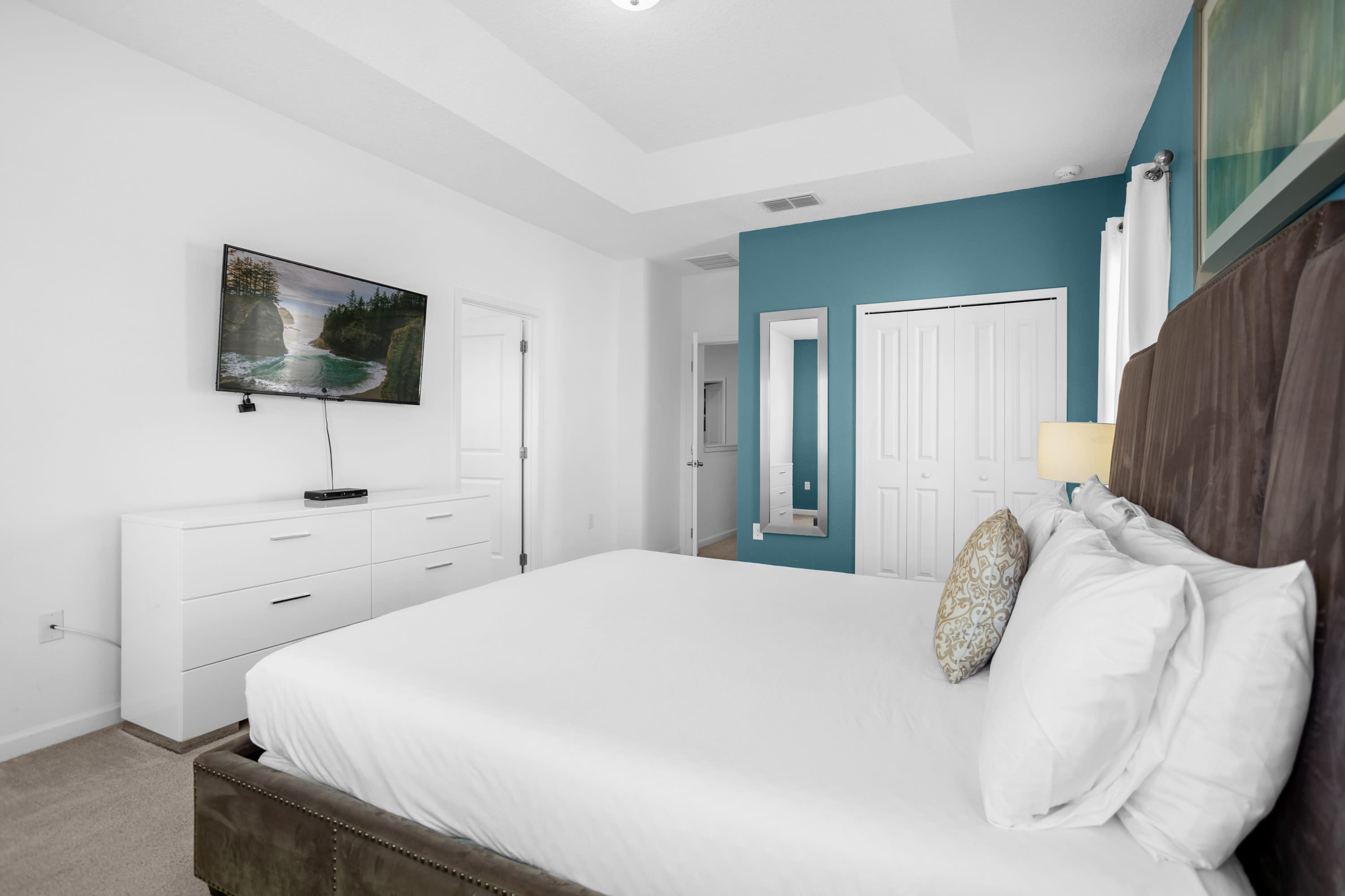 Another view of the master bedroom, showcasing the king-sized bed with its upholstered headboard, a white dresser with a wall-mounted TV, and a closet with bi-fold doors. A full-length mirror is positioned next to the closet.