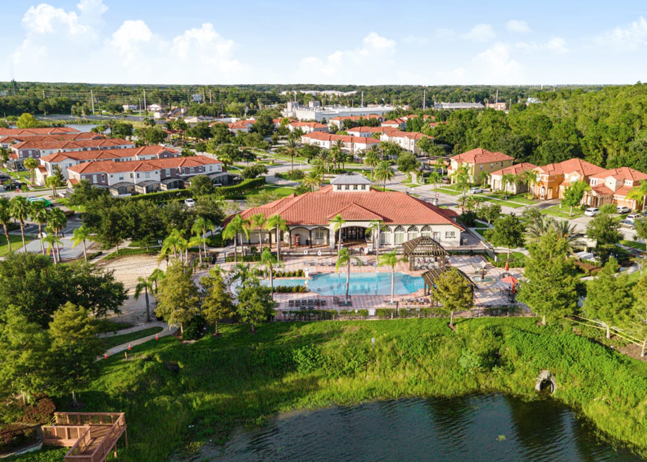 An expansive aerial view captures a residential community with numerous houses and a large clubhouse with a pool, bordered by a serene lake and lush green areas under a clear sky.