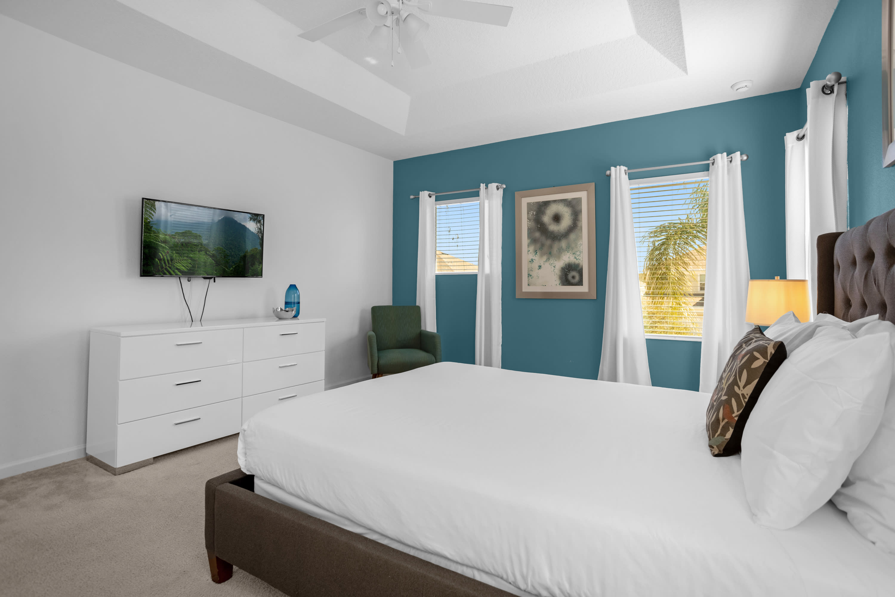 View of a bedroom with a king-sized bed, a white dresser with a wall-mounted TV, and a teal accent wall with two windows. A green accent chair is visible in the corner.