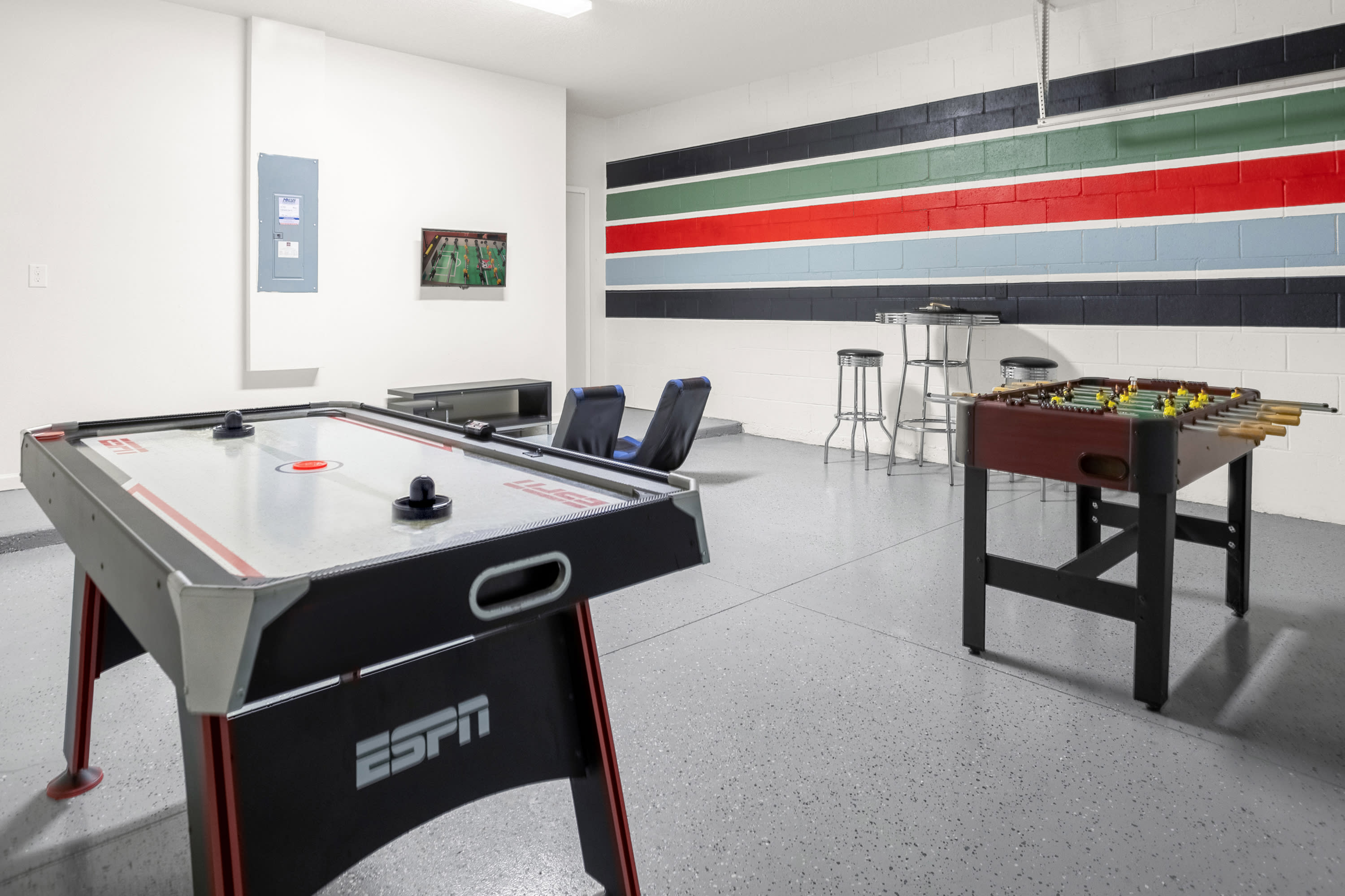 A spacious game room with white walls and a grey speckled floor features an air hockey table and a foosball table. A colorful striped accent wall adds a playful touch, with high-top tables and stools in the background.