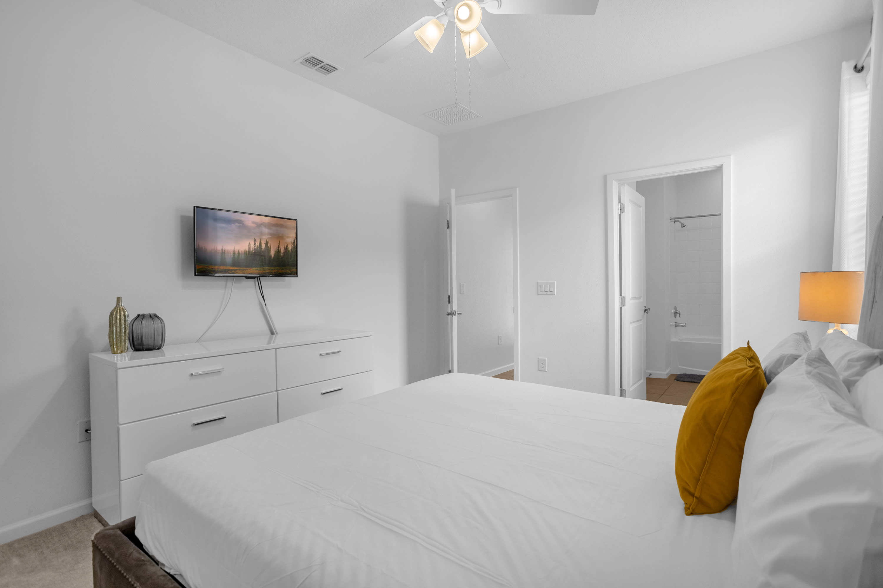 Bright white bedroom featuring a queen-sized bed with a yellow accent pillow, a white dresser with a TV on a stand, and a ceiling fan. Two doors lead to other areas, one appearing to be an en-suite bathroom.
