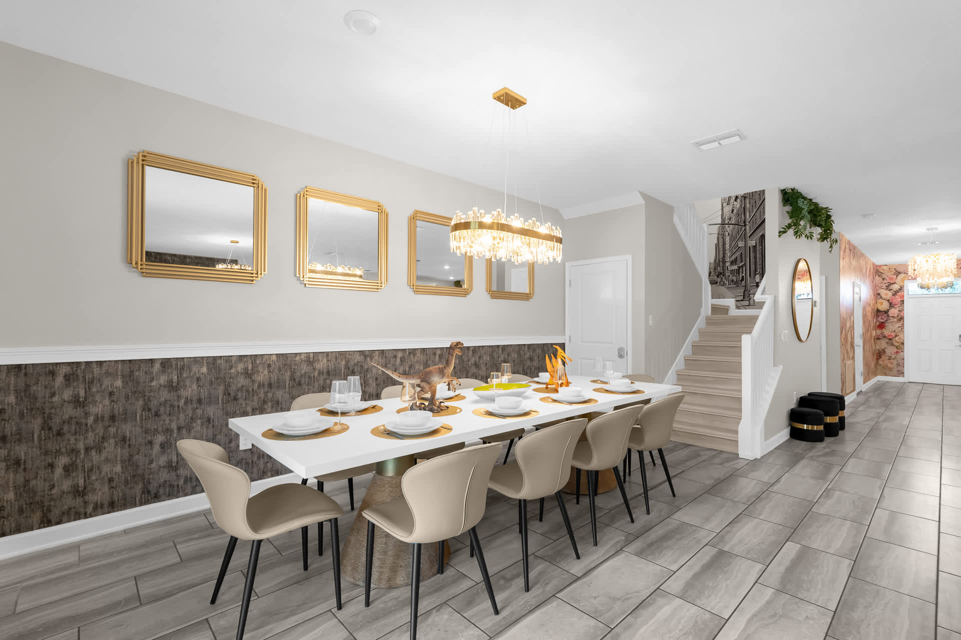 A long dining table with beige chairs is centrally placed, flanked by gold-framed mirrors and a textured wall, leading towards a staircase and front entrance.