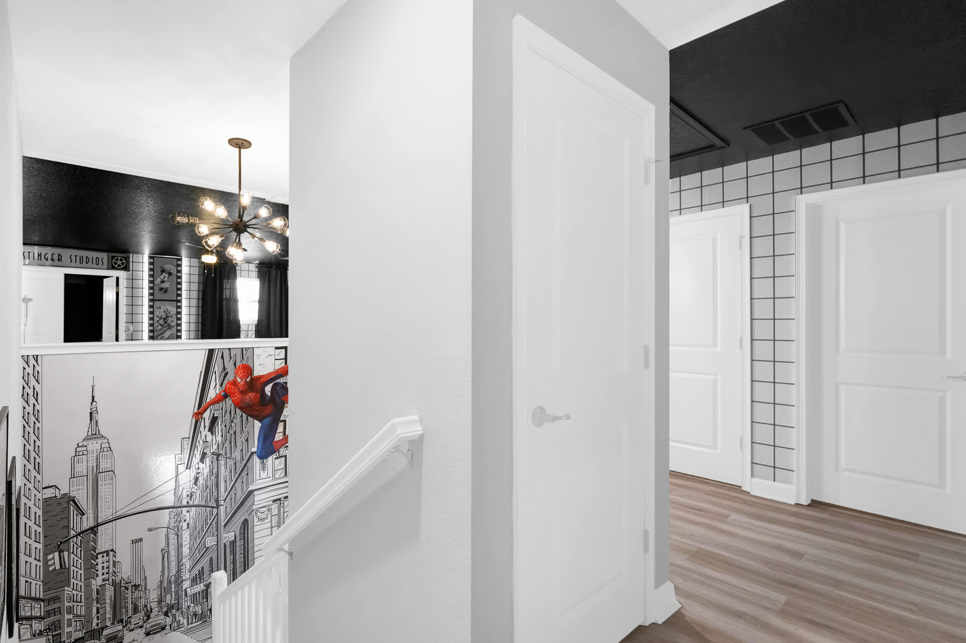 Hallway with a Spiderman mural on the wall next to a white staircase railing, leading towards a room with a black ceiling and grid-patterned walls, suggesting a themed entertainment space.