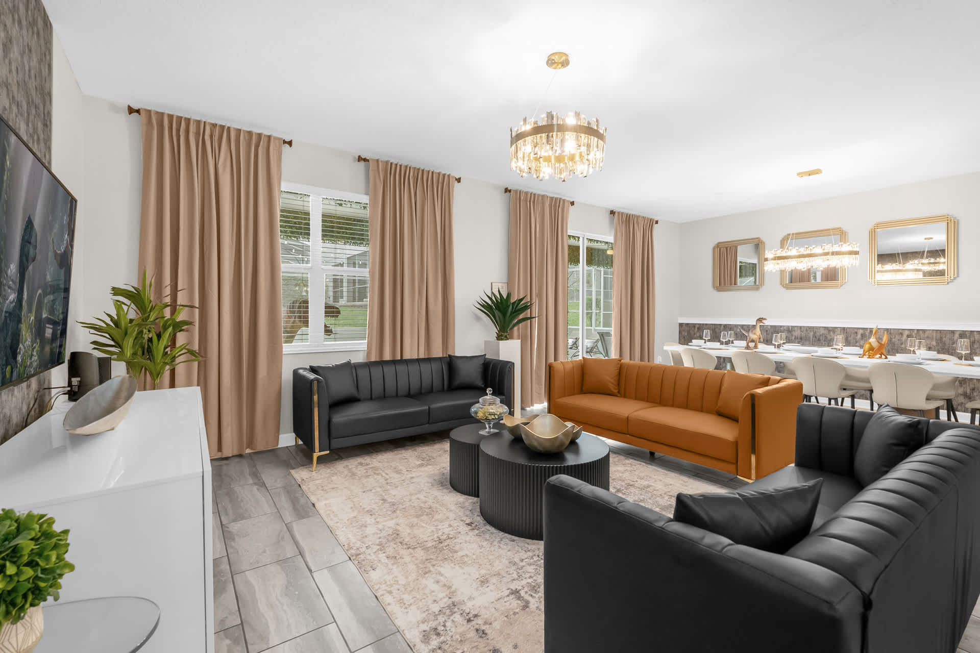A modern living room features two black sofas and one orange sofa arranged around two round coffee tables, with a large area rug and a view towards the dining area.