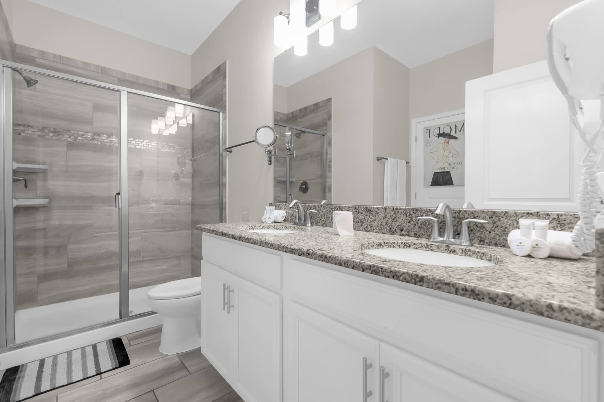 A spacious bathroom with a double vanity featuring granite countertops, white cabinets, and a large mirror. A walk-in shower with glass doors and a toilet are visible.