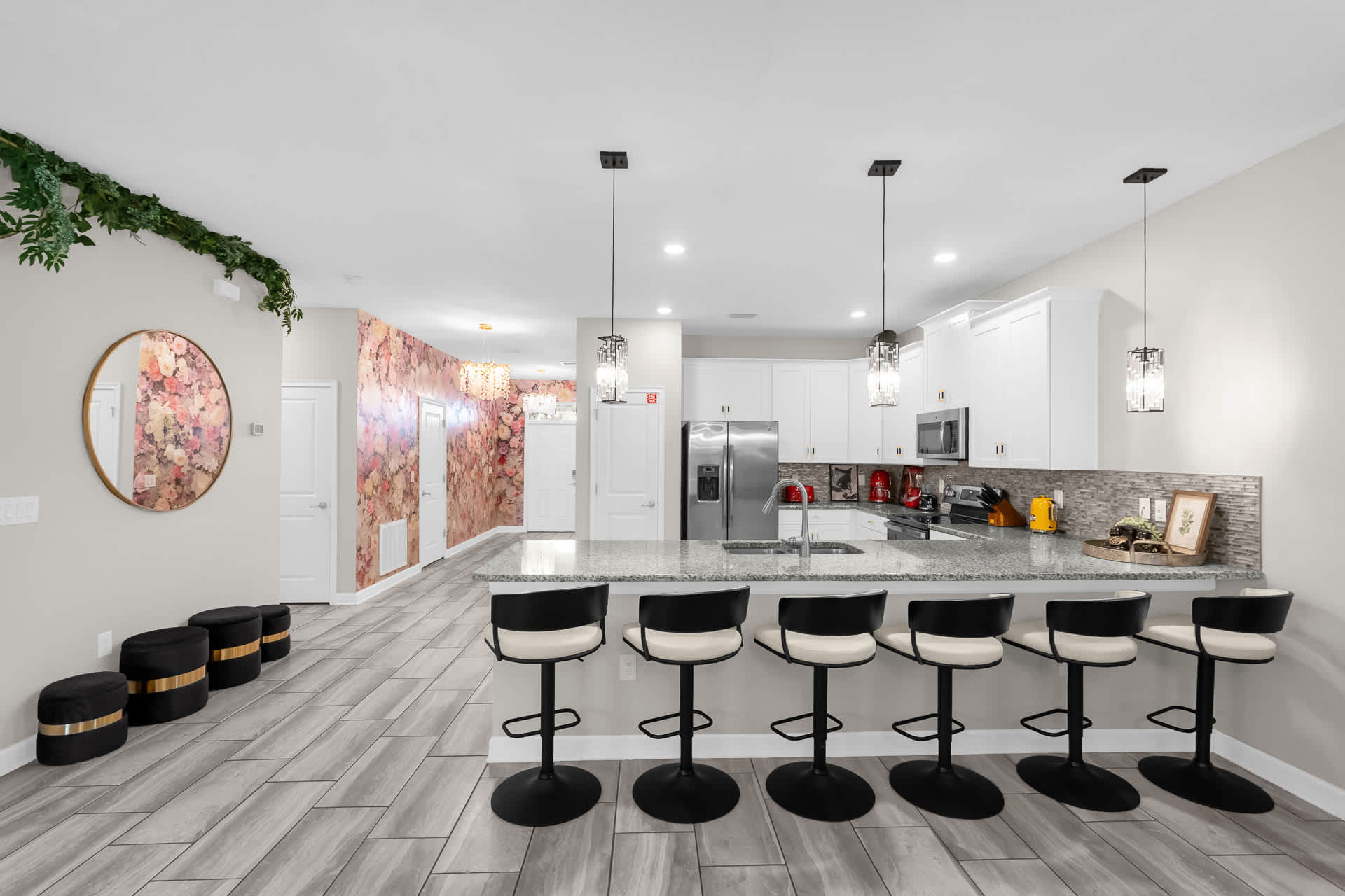Modern kitchen featuring white cabinetry, stainless steel appliances, a granite island with six black and white bar stools, and light grey tiled flooring, with a floral-patterned hallway visible in the background.