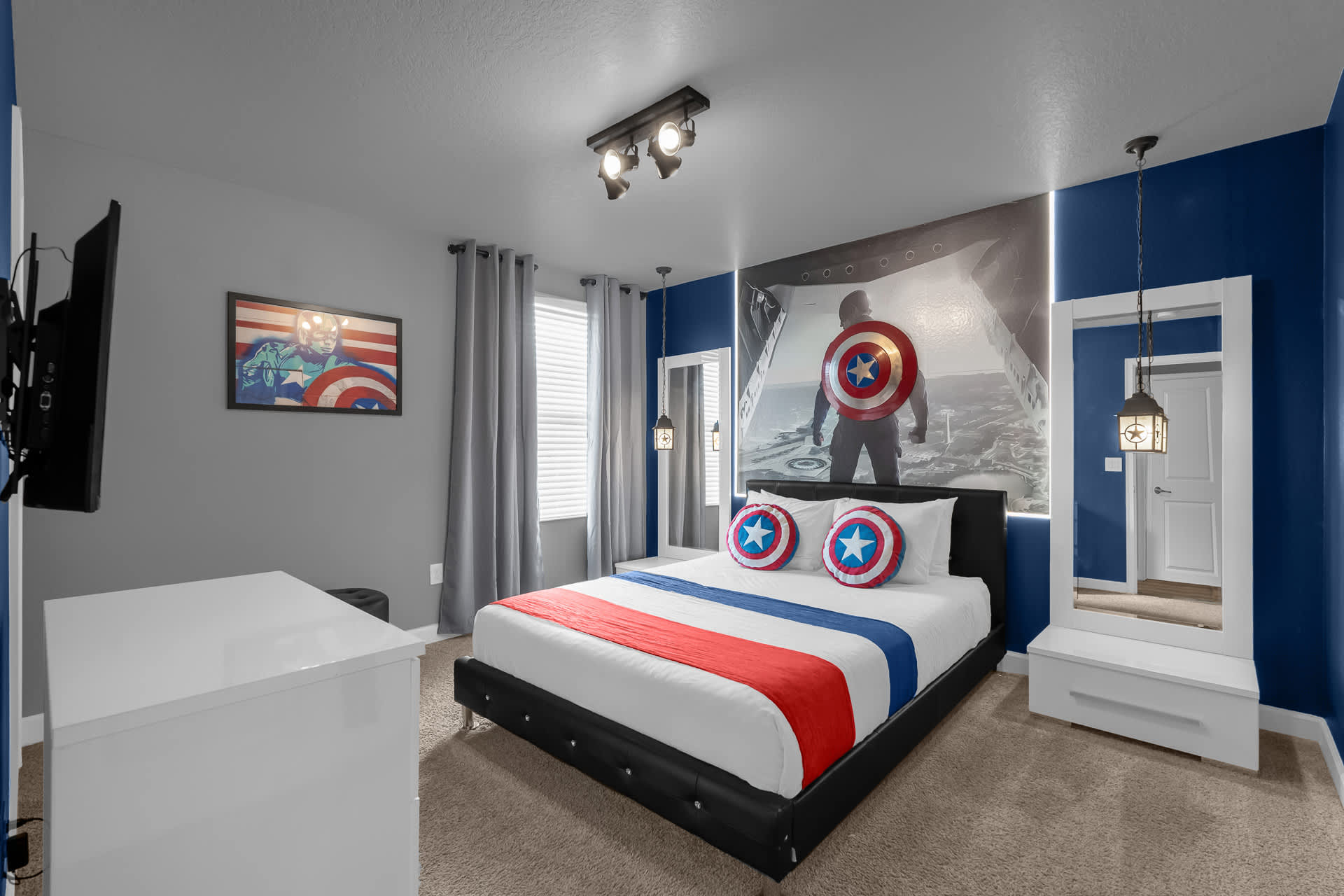 A Captain America-themed bedroom with a queen-sized bed featuring red, white, and blue bedding and shield pillows, flanked by two full-length mirrors. A large mural of Captain America covers the wall behind the bed, and a flat-screen TV is mounted on the grey wall.