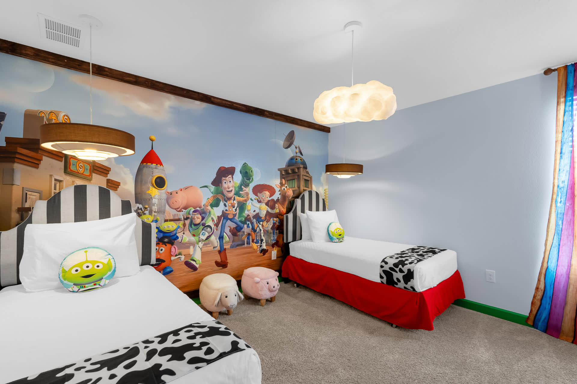 A colorful Toy Story-themed bedroom with two twin beds, each featuring a striped headboard and alien-themed pillows. A large mural of Toy Story characters covers the wall, and the room is decorated with cloud-shaped pendant lights.