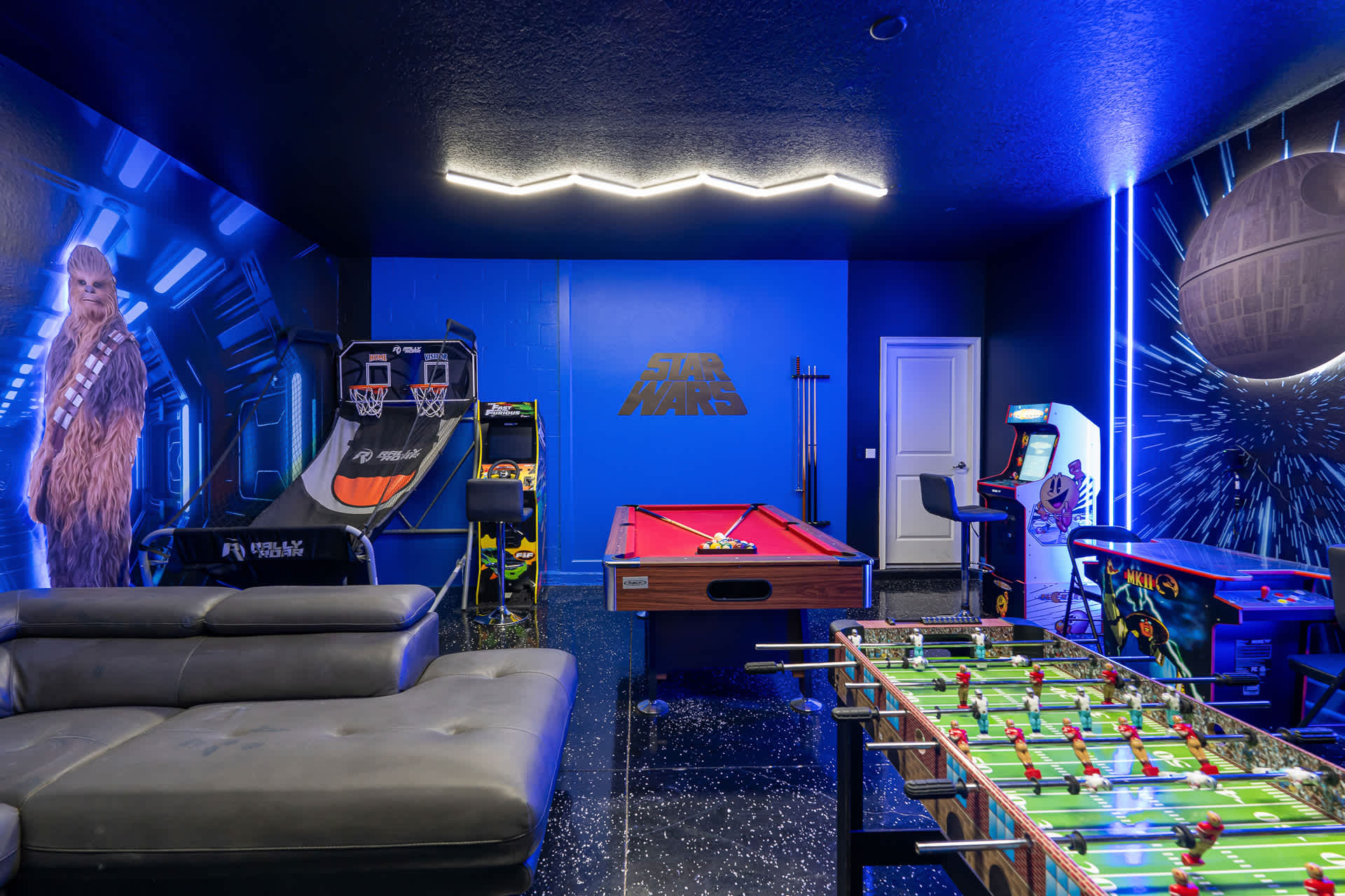 A wide view of the Star Wars-themed game room, showcasing a pool table, foosball, basketball arcade game, and other arcade machines under bright neon strip lighting.
