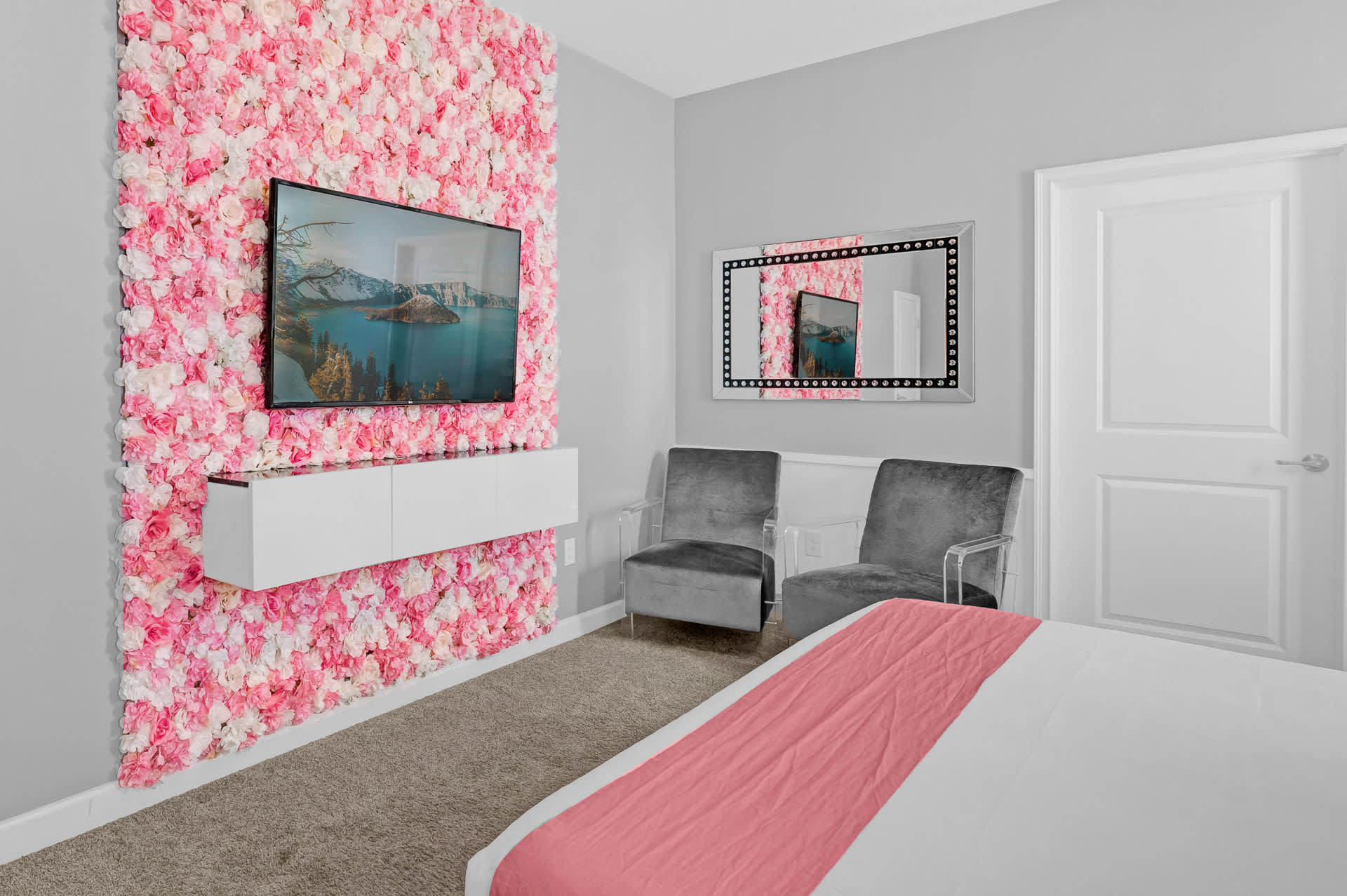 Chic bedroom featuring a pink floral accent wall with a mounted TV, two grey velvet armchairs with clear acrylic frames, and a large mirrored dresser, all set against light grey walls and carpeted flooring.