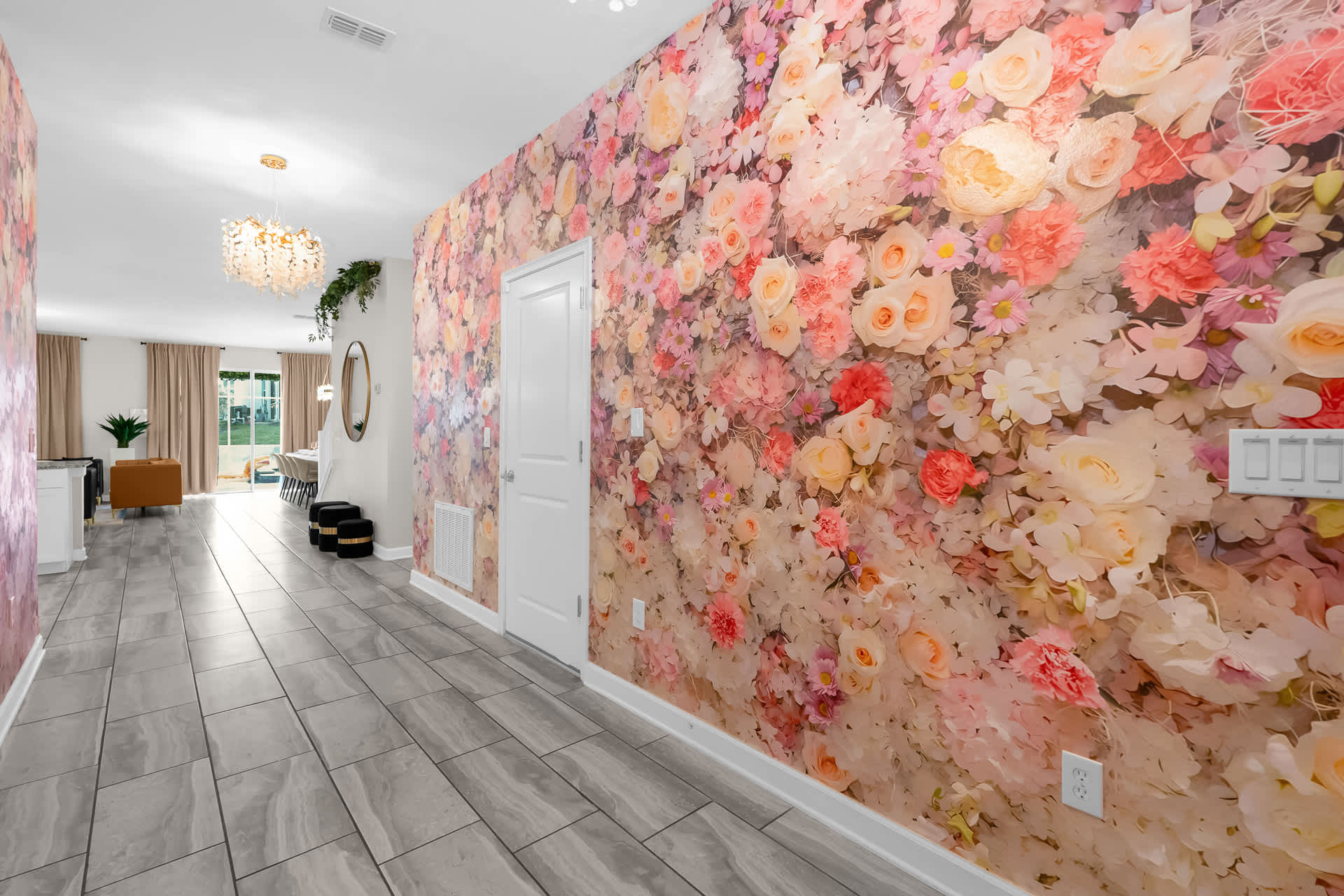 A bright hallway with light grey tiled flooring and a striking accent wall covered in a large floral wallpaper pattern featuring pink, white, and orange flowers. A white door is visible on the right, and a glimpse of a dining area is seen in the background.
