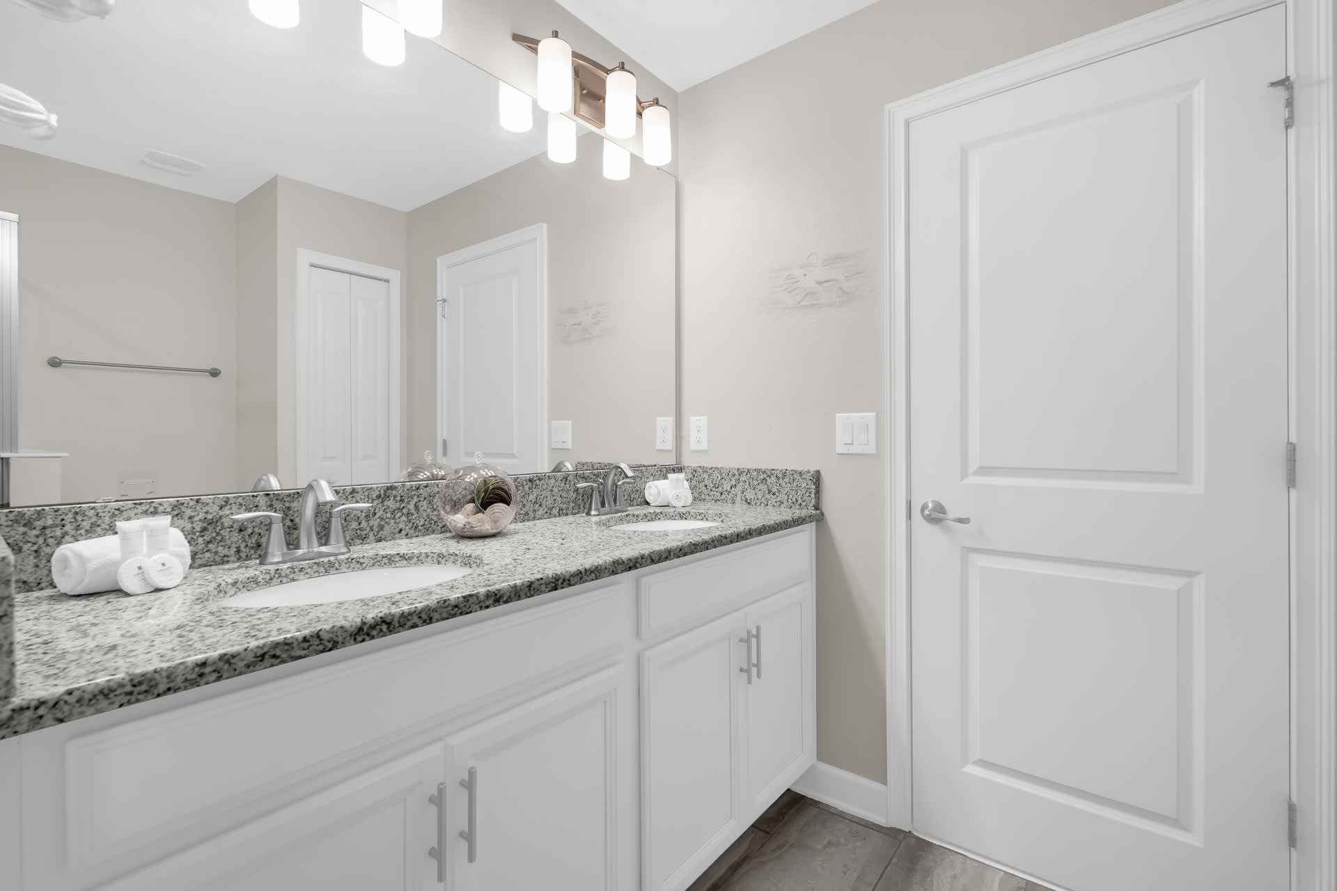 A clean bathroom featuring a double vanity with granite countertops, white shaker-style cabinets, and a large mirror. The floor is tiled with light gray wood-look planks.
