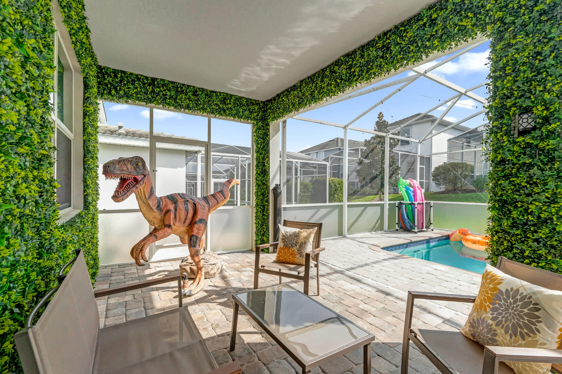 A screened-in patio features a large dinosaur statue, outdoor seating with a coffee table, and artificial green wall coverings, overlooking a private pool area.