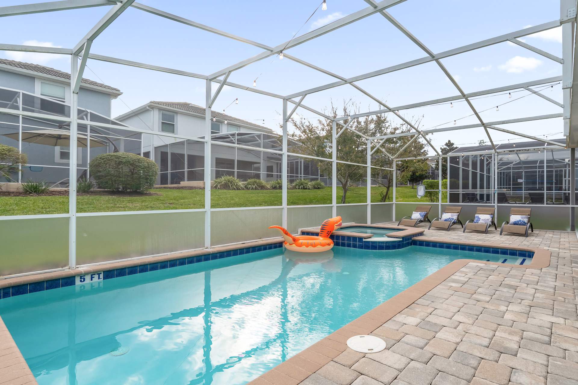 A private screened swimming pool area features a rectangular pool with an attached hot tub, surrounded by light-colored pavers and a few lounge chairs. The enclosure offers views of neighboring houses and a grassy backyard.
