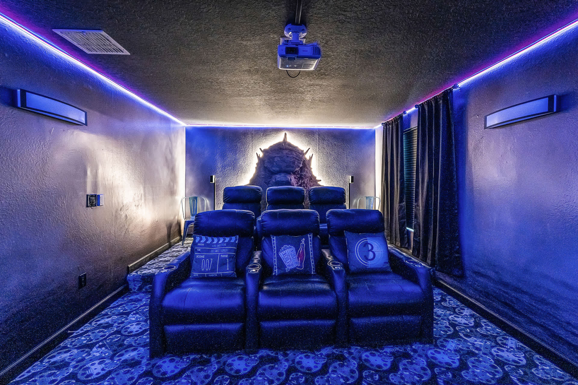 The media room features multiple rows of plush reclining theater seats, a projector, and a unique illuminated wall sculpture behind the back row, all under blue ambient lighting.