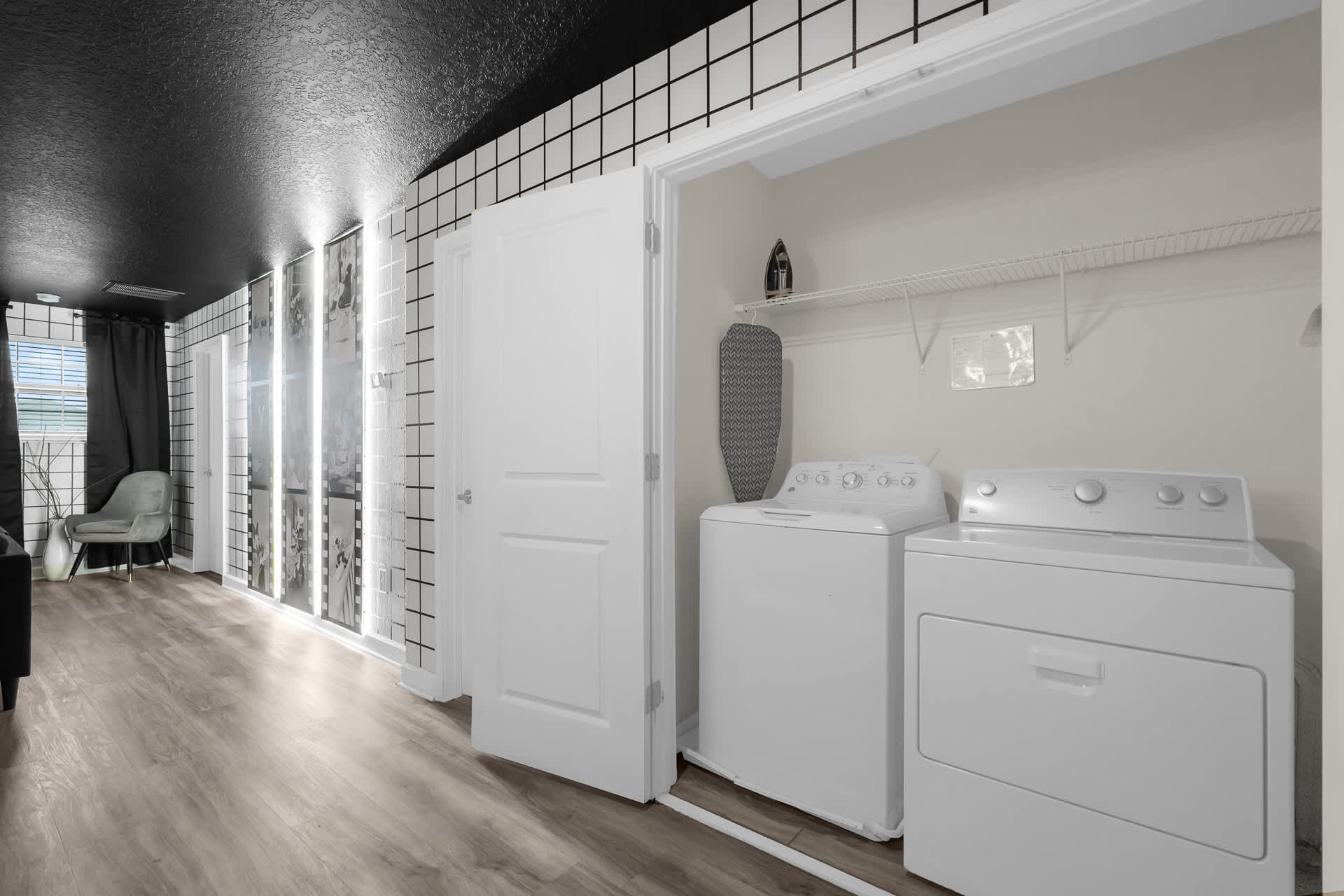 A dedicated laundry area featuring a white top-loading washing machine and a matching dryer, neatly tucked into a closet space. An ironing board and iron are stored on a shelf above the appliances.