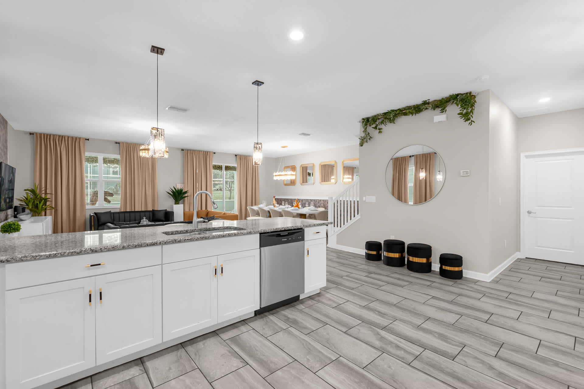 Open-concept kitchen with a large granite island and white cabinetry, overlooking a spacious living area with light-colored curtains and a dining space, all featuring light grey tiled flooring.