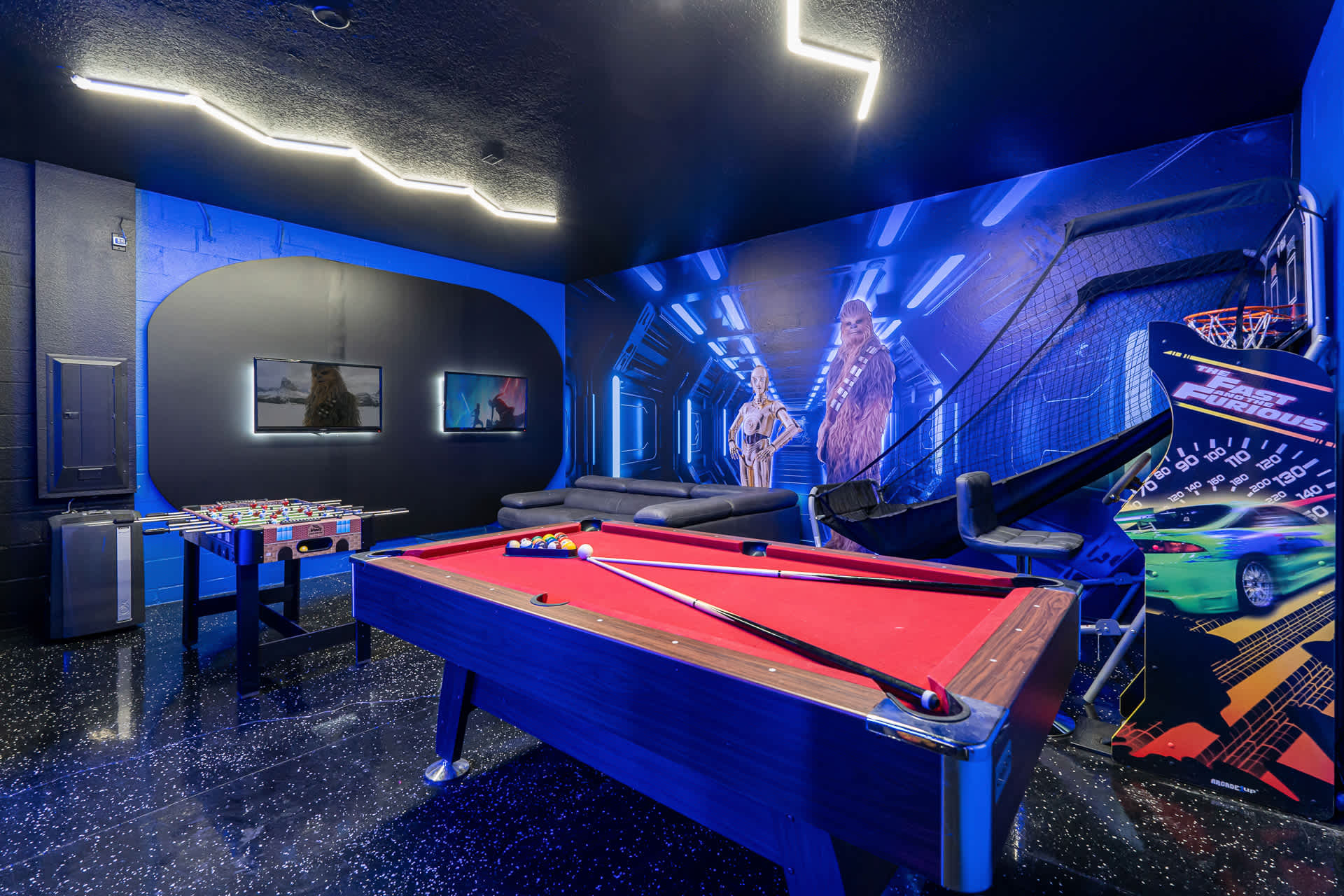 A vibrant game room features a red felt pool table, foosball table, and arcade games, all set against dark blue walls with Star Wars-themed murals and neon lighting.