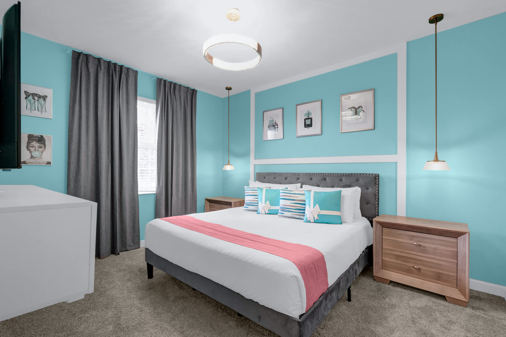 A vibrant bedroom with bright blue accent walls and a queen-sized bed featuring a gray upholstered headboard, white bedding, and colorful throw pillows. Two wooden nightstands with pendant lights flank the bed.