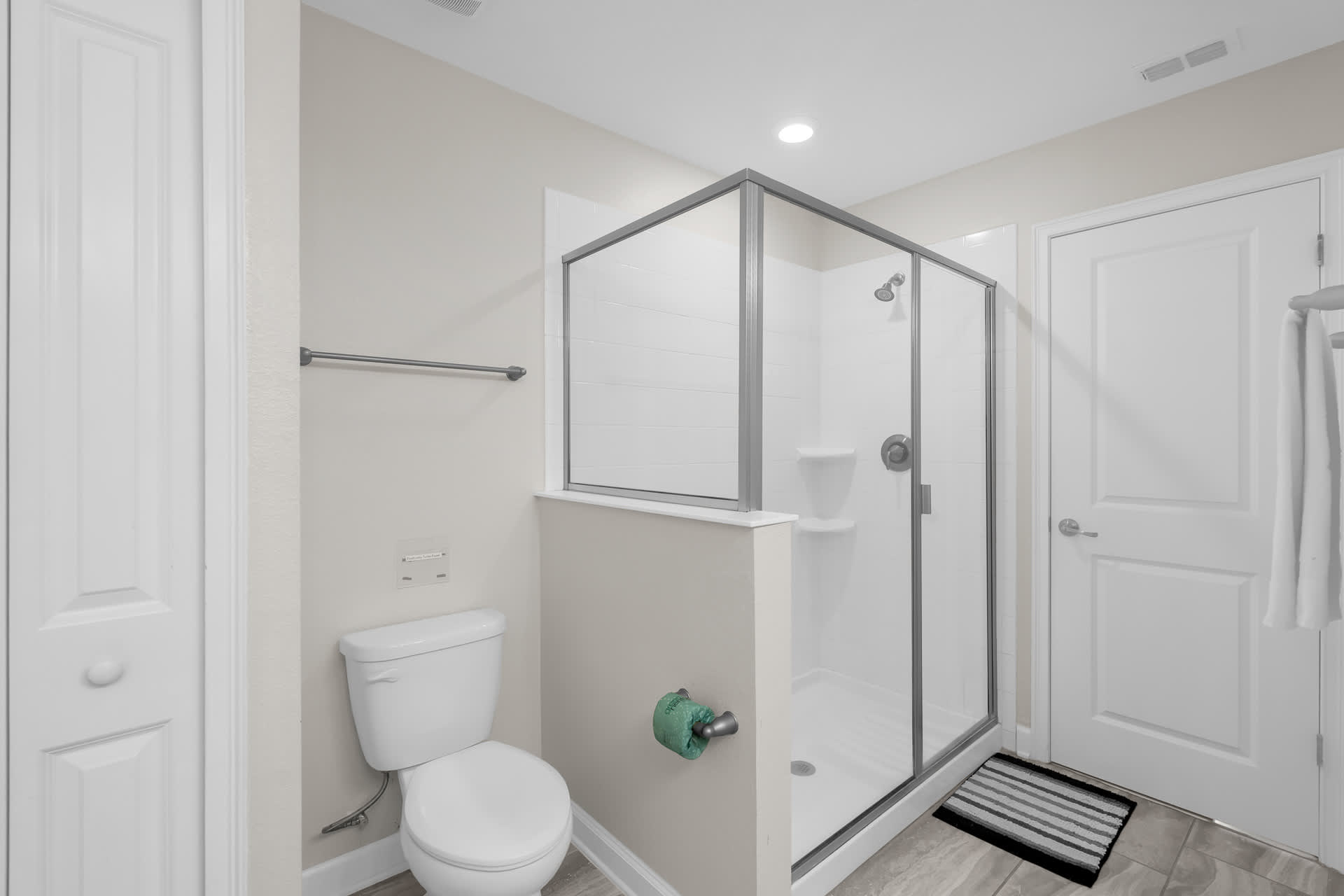 A modern bathroom featuring a white toilet, a glass-enclosed walk-in shower with white subway tiles, and light beige walls. A towel bar is mounted above the toilet.