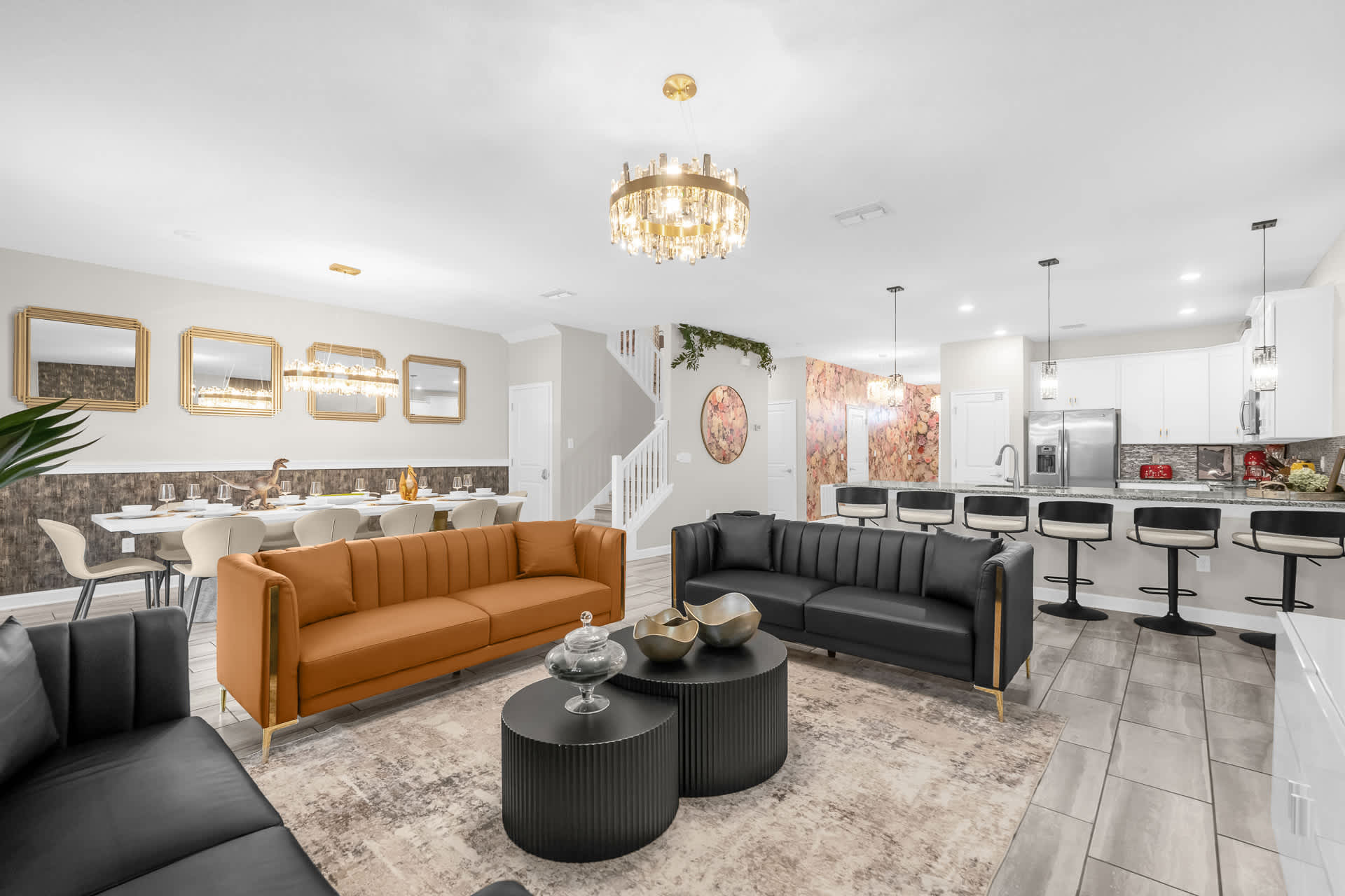 An open-concept living space features a modern design with two large sofas, one orange and one black, facing a coffee table on a patterned rug. A dining area with a long white table and a kitchen with a breakfast bar and white cabinetry are visible.