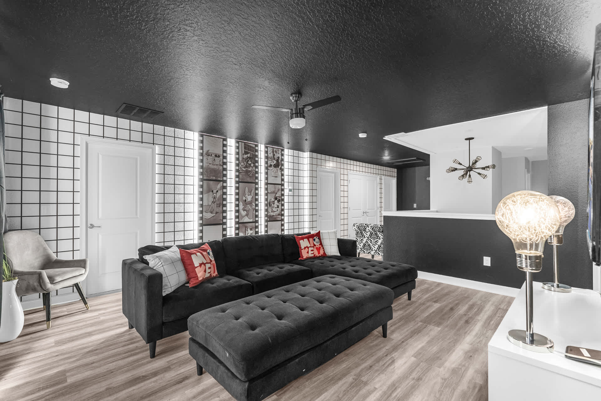 Themed media room featuring a large black sectional sofa with ottomans, a black textured ceiling, and white walls with a black grid pattern, illuminated by a ceiling fan and decorative lamps.