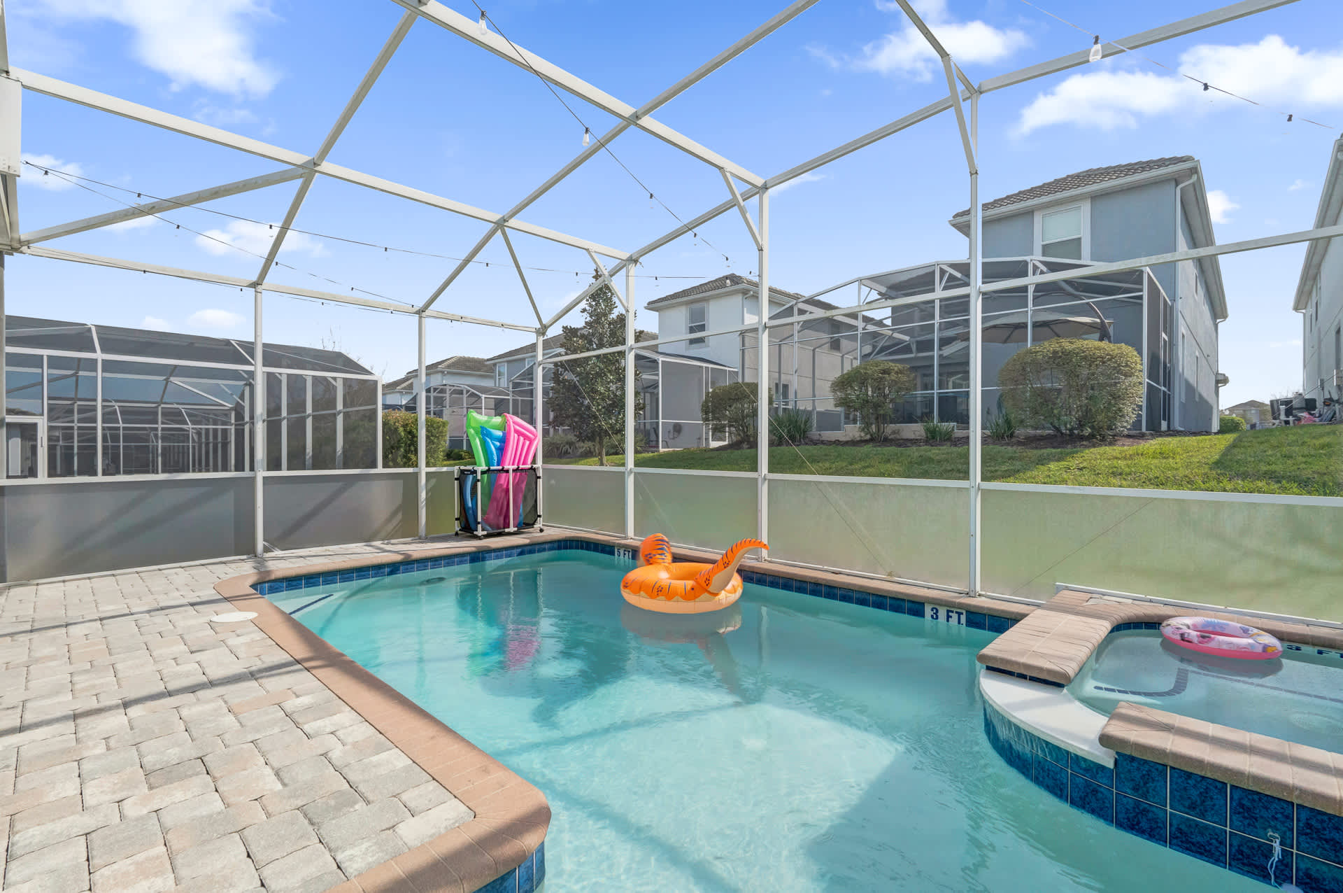 A private screened swimming pool area showcases a rectangular pool with a shallow entry and an attached hot tub, surrounded by light-colored pavers. Pool floats and additional lounge chairs are visible, with neighboring houses in the background.