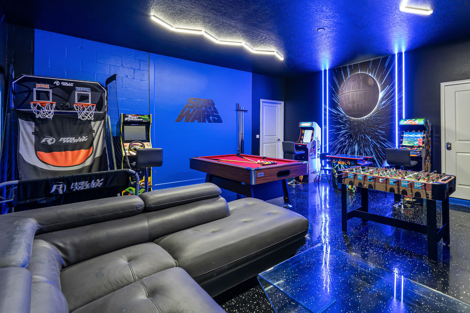 A vibrant Star Wars-themed game room features electric blue walls, neon strip lighting, a red-felt pool table, arcade machines, and a foosball table, all set on a glossy black floor with white speckles. A large sectional sofa provides seating.