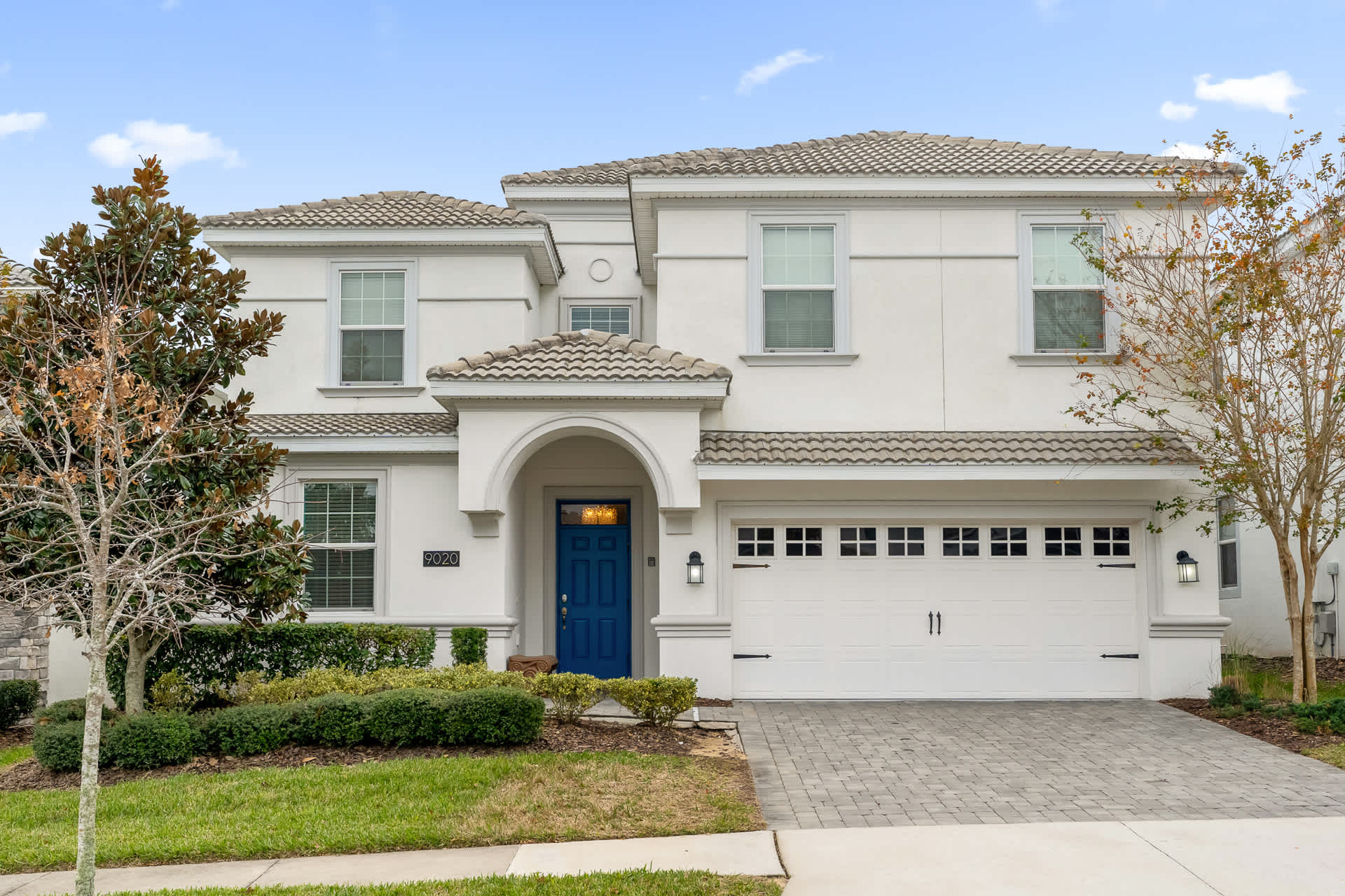 The front exterior of a large, two-story white house features a prominent blue front door under an arched entryway, a two-car garage, and a paved driveway. Landscaped bushes and small trees adorn the front yard.