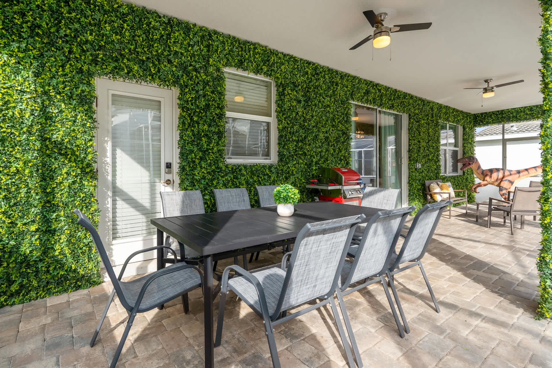 A spacious covered patio features an outdoor dining table with eight chairs, a BBQ grill, and artificial green wall coverings, with a view of the pool area.