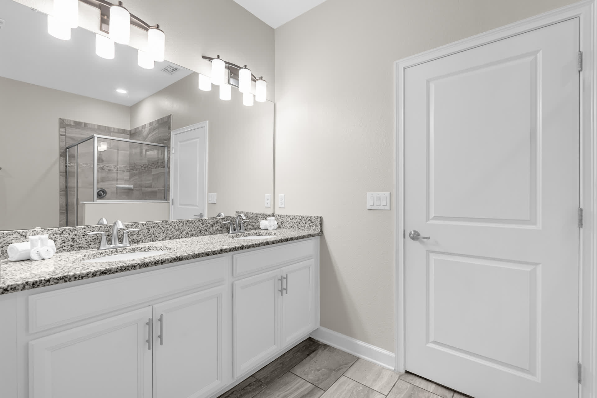 A spacious bathroom with a double vanity featuring granite countertops, two undermount sinks, and white shaker-style cabinets. A large mirror spans the width of the vanity, reflecting a walk-in shower in the background.