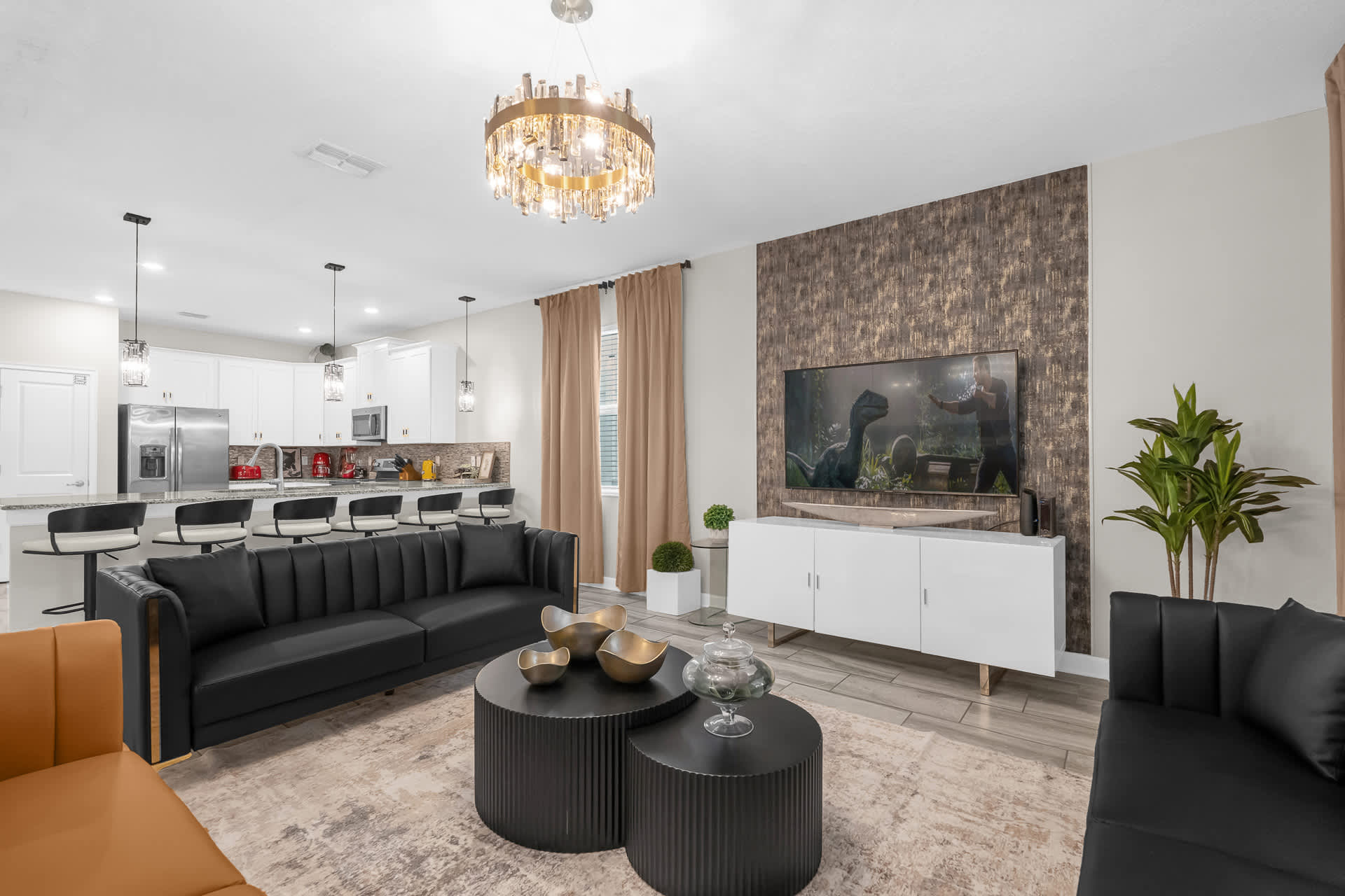 An open-concept living area features black and orange sofas, round coffee tables, and a large TV mounted on a textured accent wall, with a modern kitchen visible in the background.