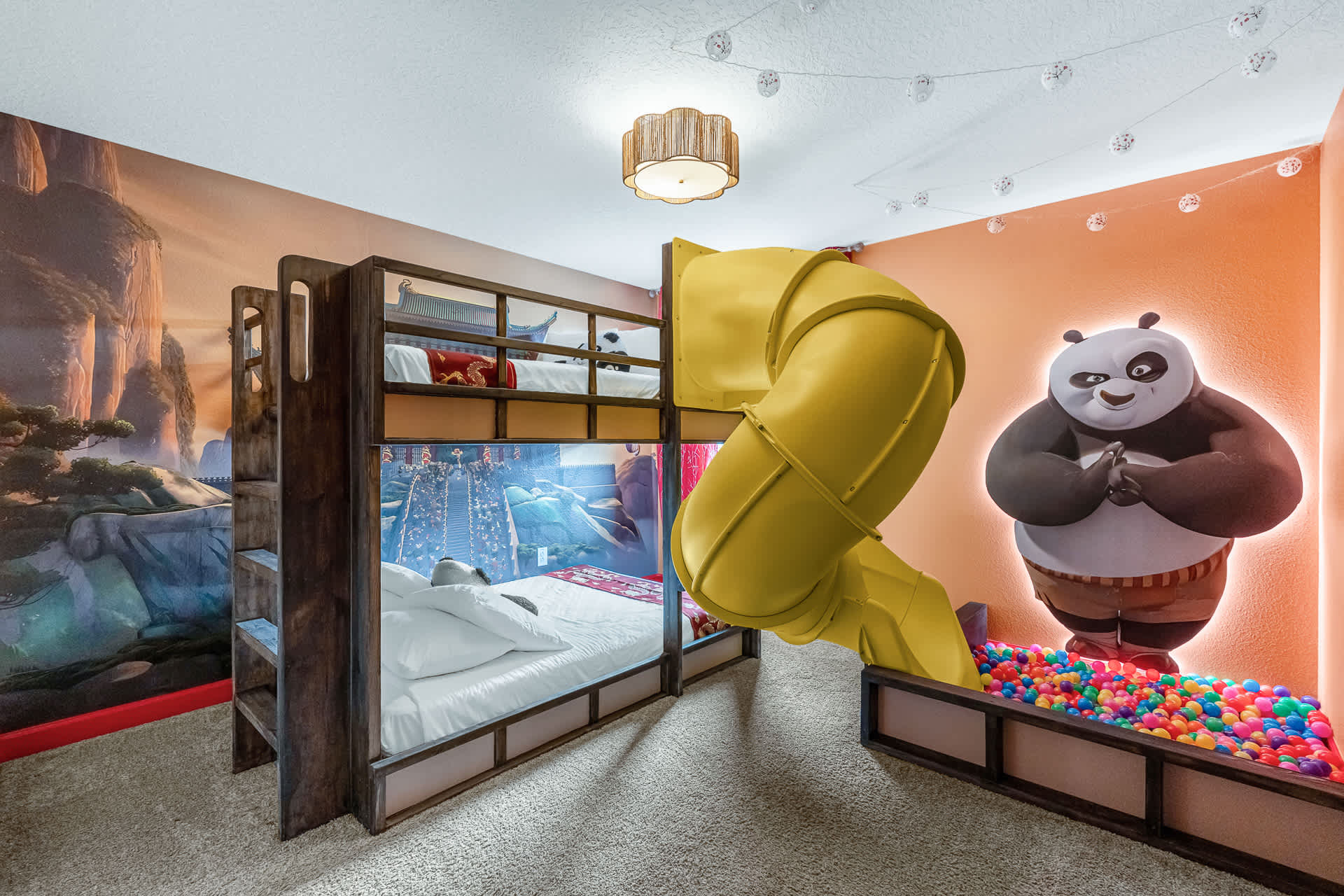 A vibrant Kung Fu Panda-themed kids' bedroom featuring a wooden bunk bed with a yellow spiral slide leading into a ball pit. A large illuminated mural of Po the panda adorns the orange wall, creating a playful atmosphere.