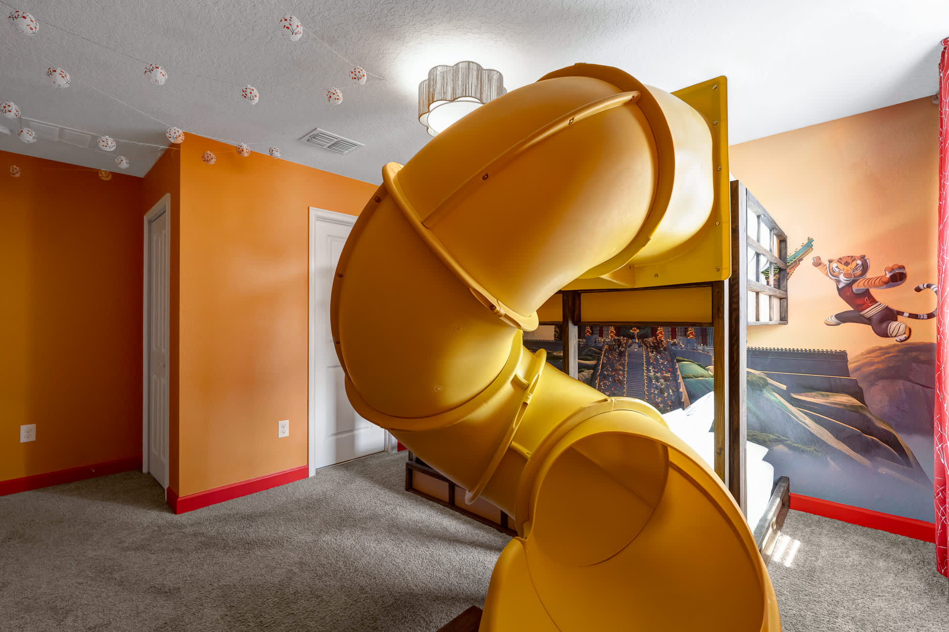 A close-up view of the yellow spiral slide in the Kung Fu Panda-themed bedroom, extending from the top bunk down to a ball pit. The room features orange walls and decorative string lights on the ceiling.