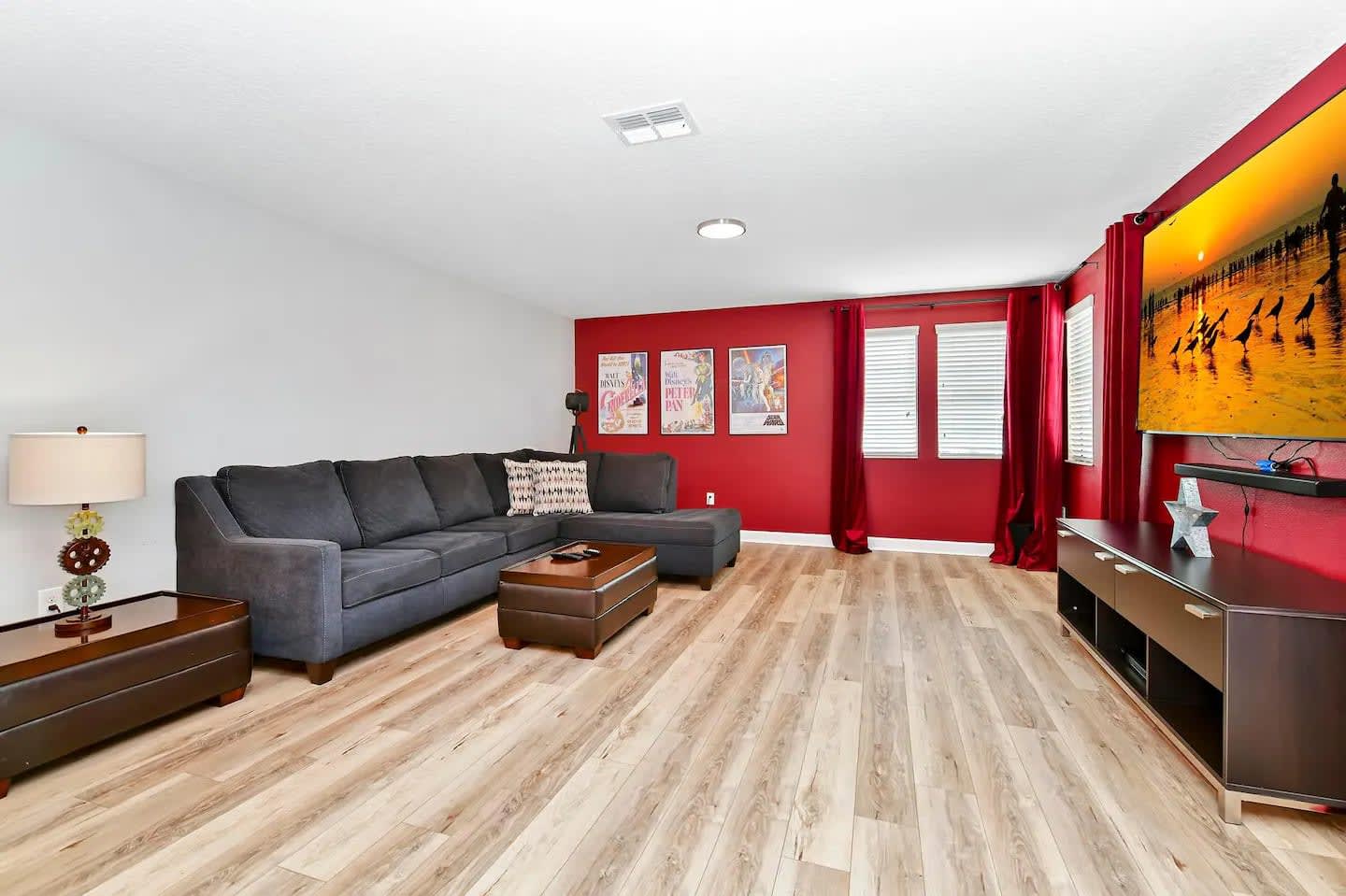A spacious living area with light wood-look flooring and a striking red accent wall, featuring a large dark gray sectional sofa and a brown ottoman. A wall-mounted flat-screen TV is visible on the right, with decorative posters on the red wall.