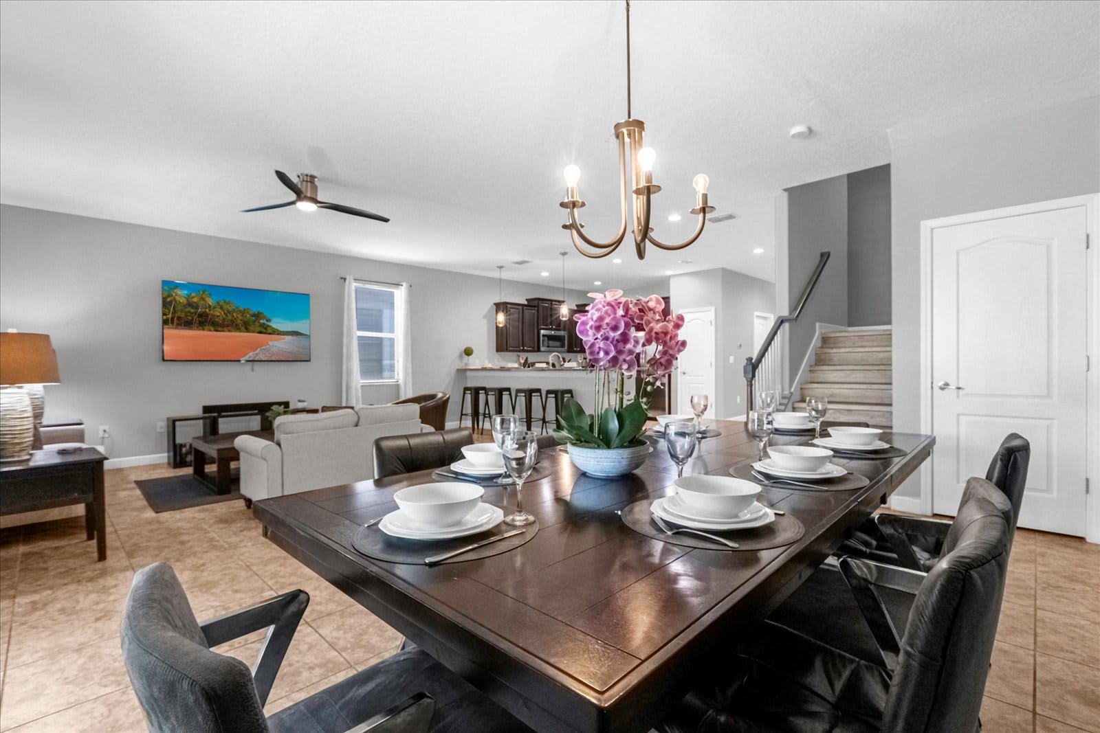 Elegant dining area with a dark wood rectangular table set for eight, adorned with a floral centerpiece and a bronze chandelier. The space opens to a living room with a TV and a kitchen bar in the background.