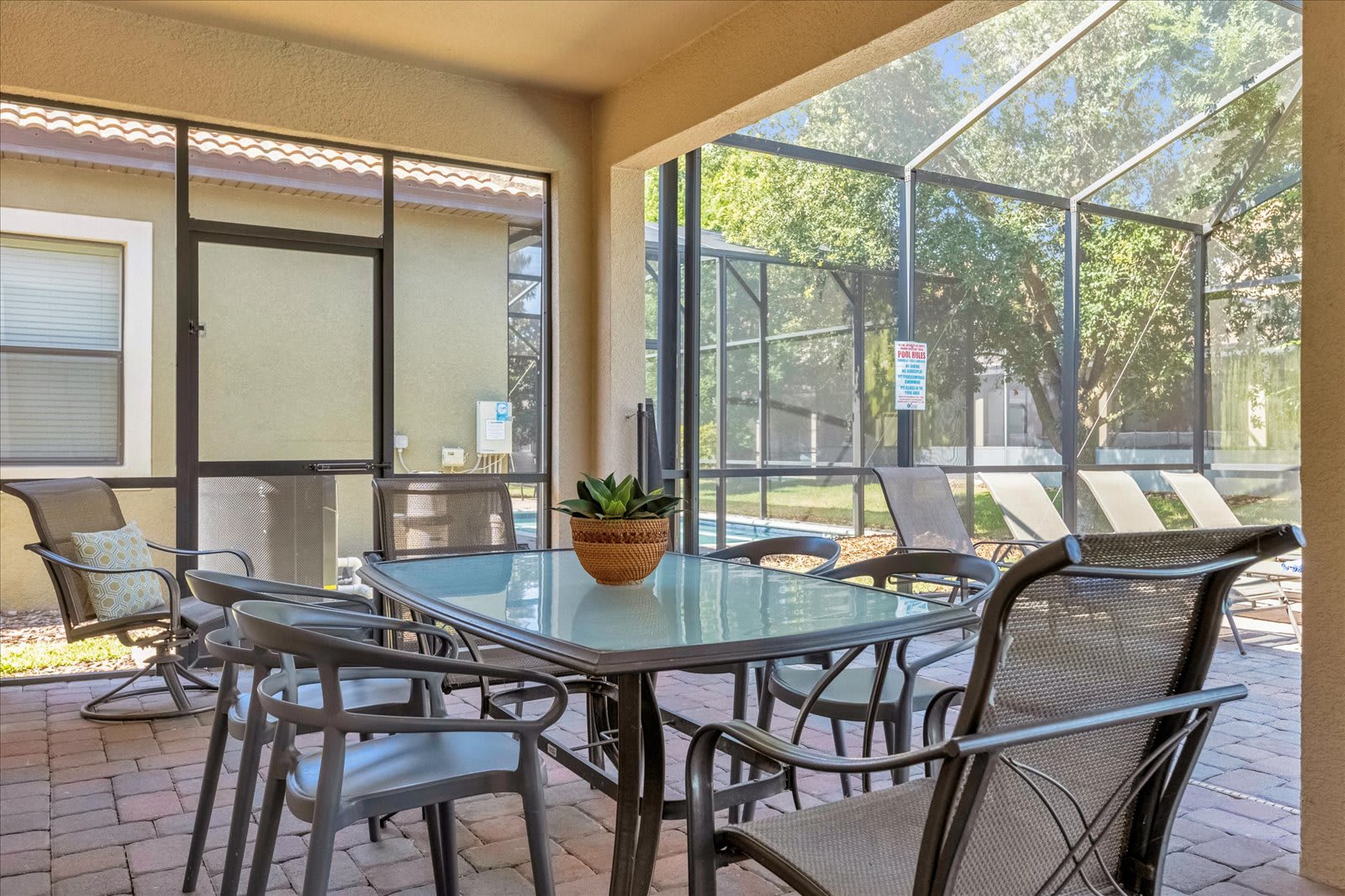A covered outdoor patio area with brick pavers, featuring a glass-top dining table surrounded by six chairs. The area is enclosed by a screen, offering views of the private pool and lush greenery beyond.