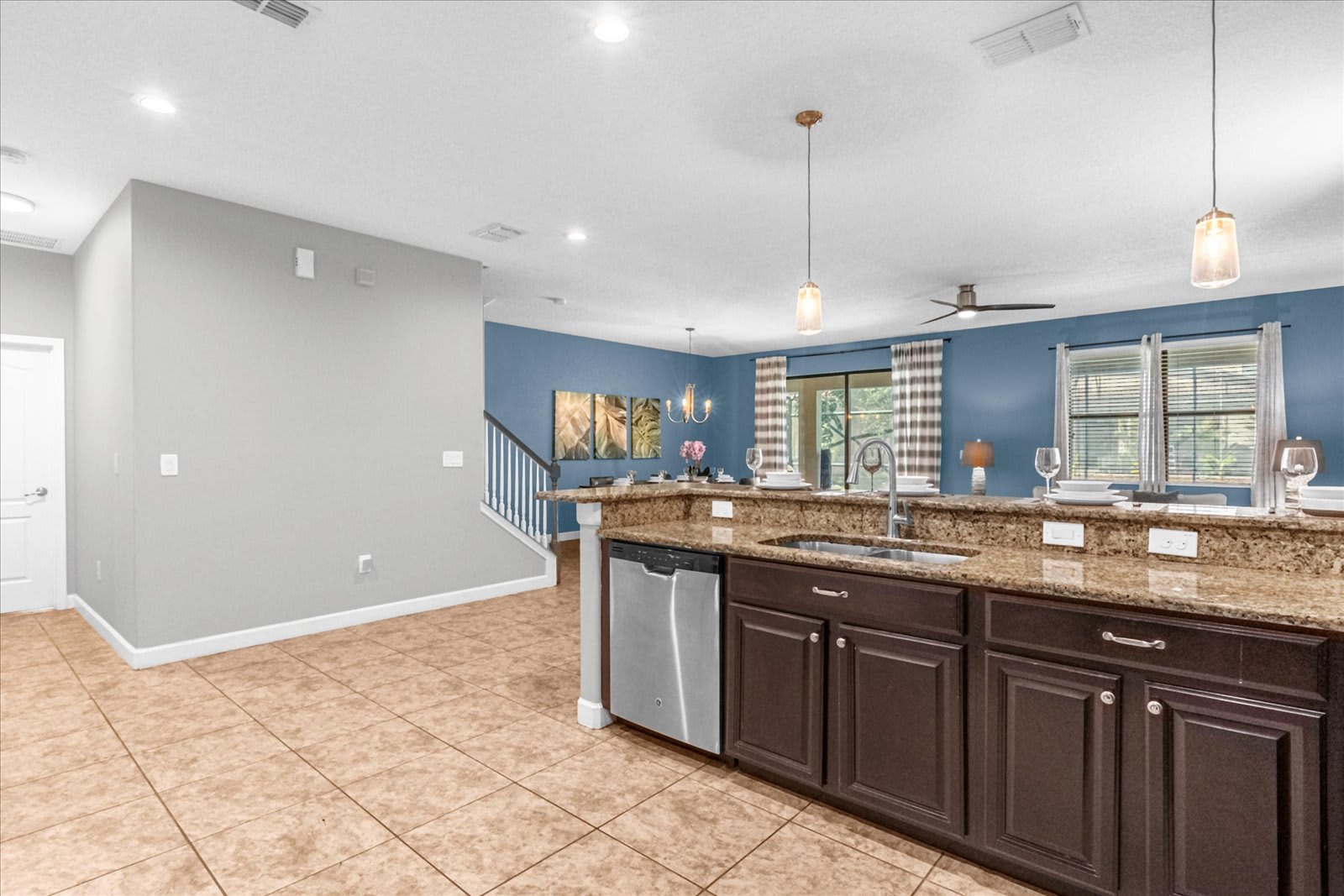 Wide shot of the kitchen and adjacent areas, showing the kitchen bar with dark cabinets and granite countertops, a stainless steel dishwasher, and pendant lighting. The open layout connects to the living and dining spaces.