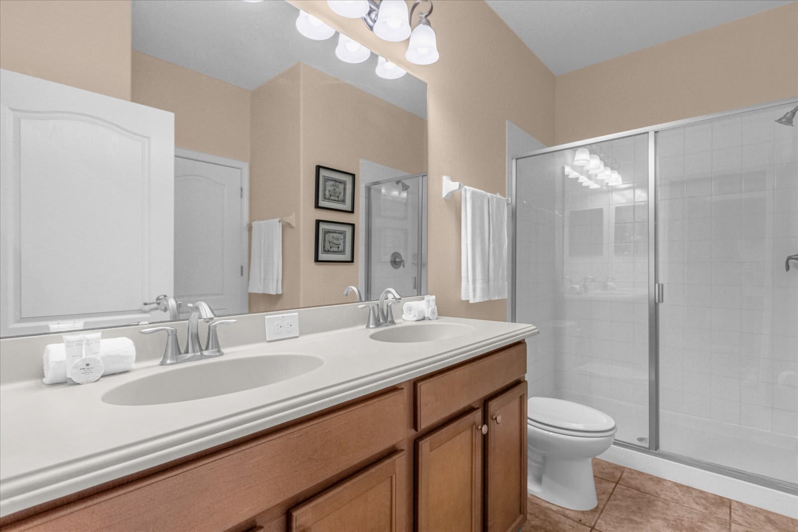 Modern bathroom with light beige walls and brown tiled flooring, featuring a double vanity with a light wood cabinet and white countertop. A large mirror reflects a walk-in shower with a glass enclosure and white tiled walls.