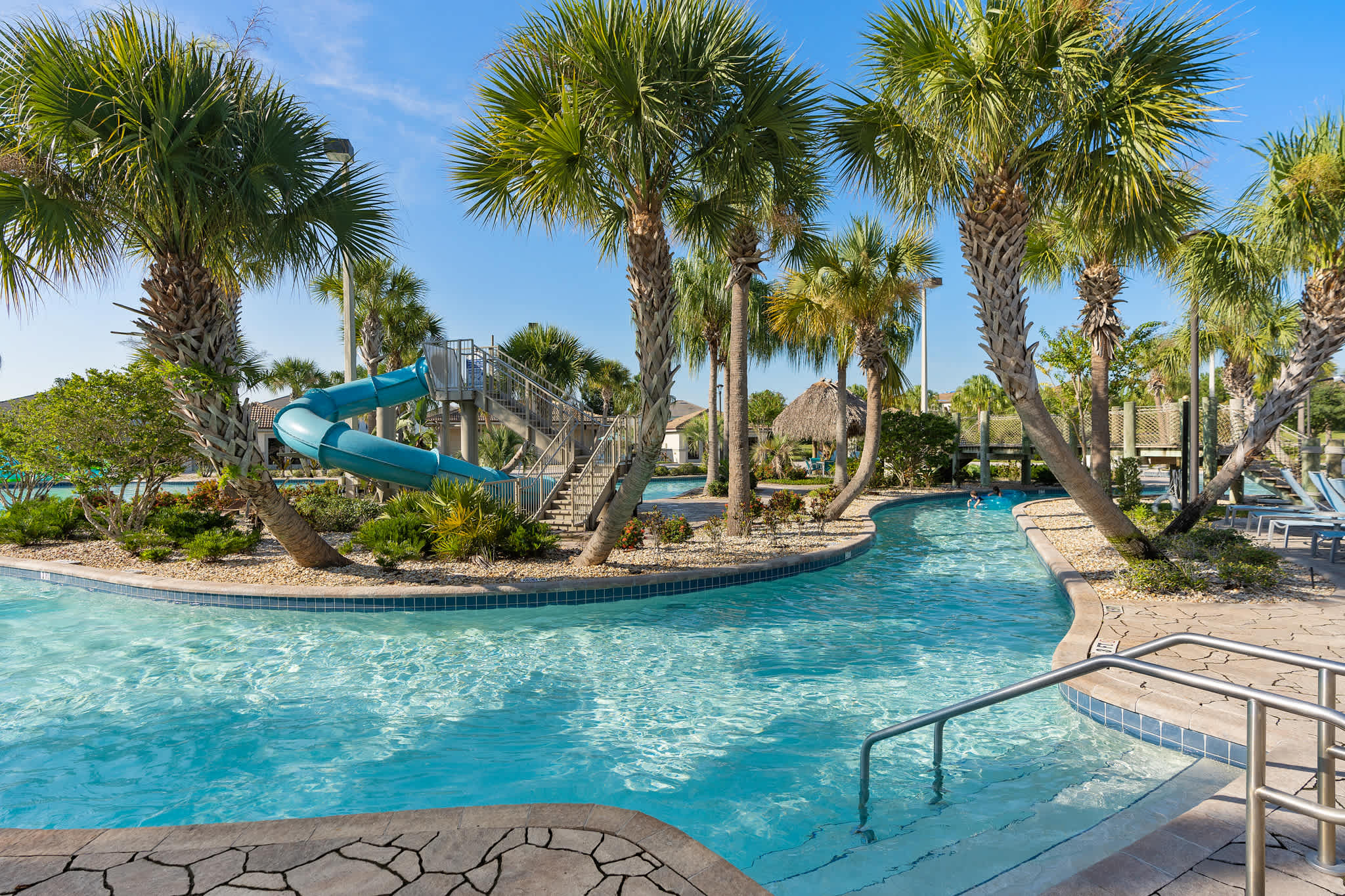 A winding lazy river at a communal pool area, surrounded by lush palm trees and tropical landscaping, with a blue water slide visible in the background.