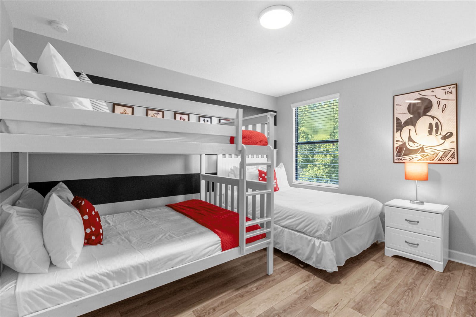 Mickey Mouse themed bedroom with a white bunk bed featuring red bedding, a single bed with white bedding, and a large Mickey Mouse pop art poster on the wall, all on light wood-look flooring.