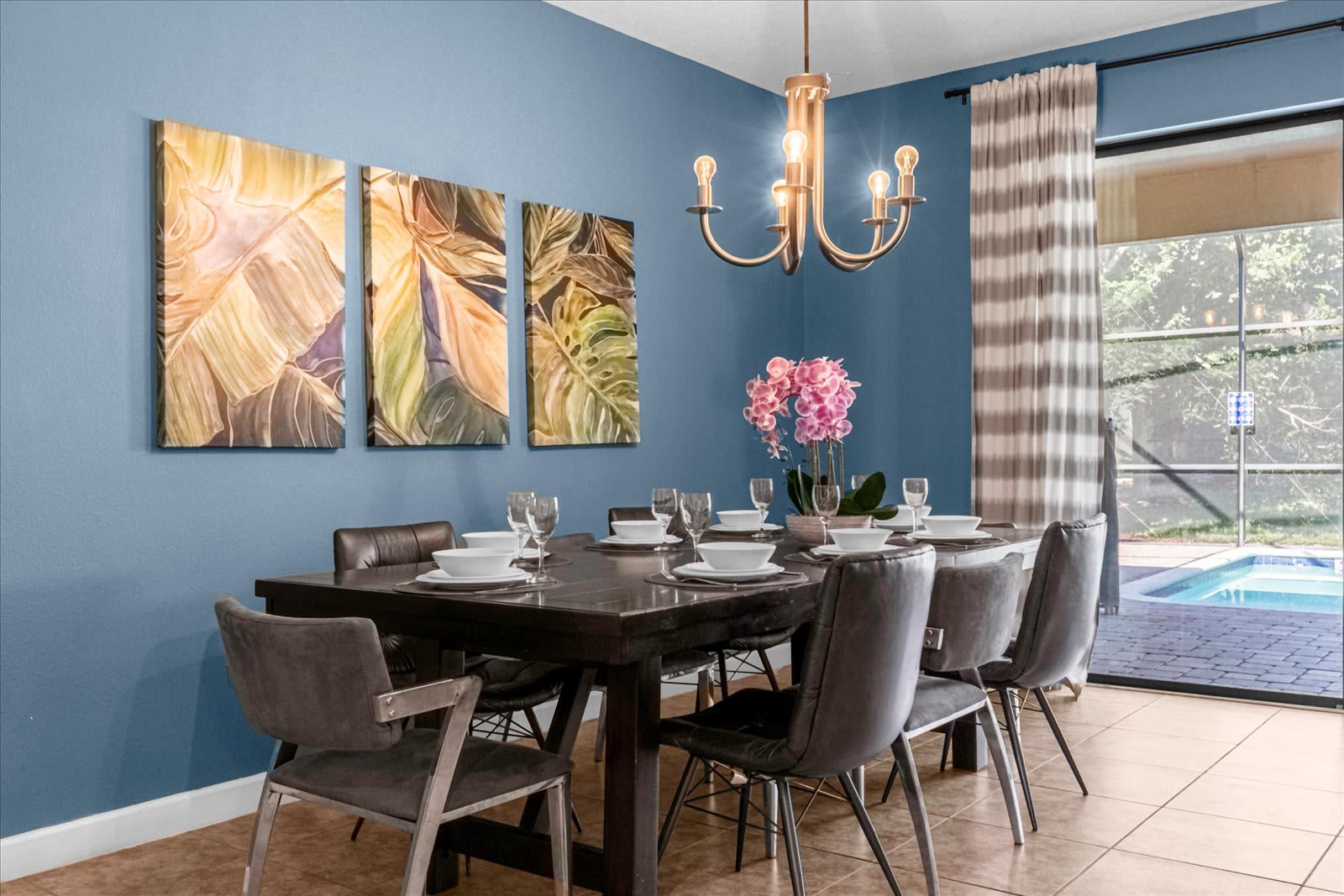 Bright dining room with a blue accent wall adorned with three botanical art pieces, featuring a dark wood table set for eight and a bronze chandelier. Large sliding glass doors lead to an outdoor pool area.