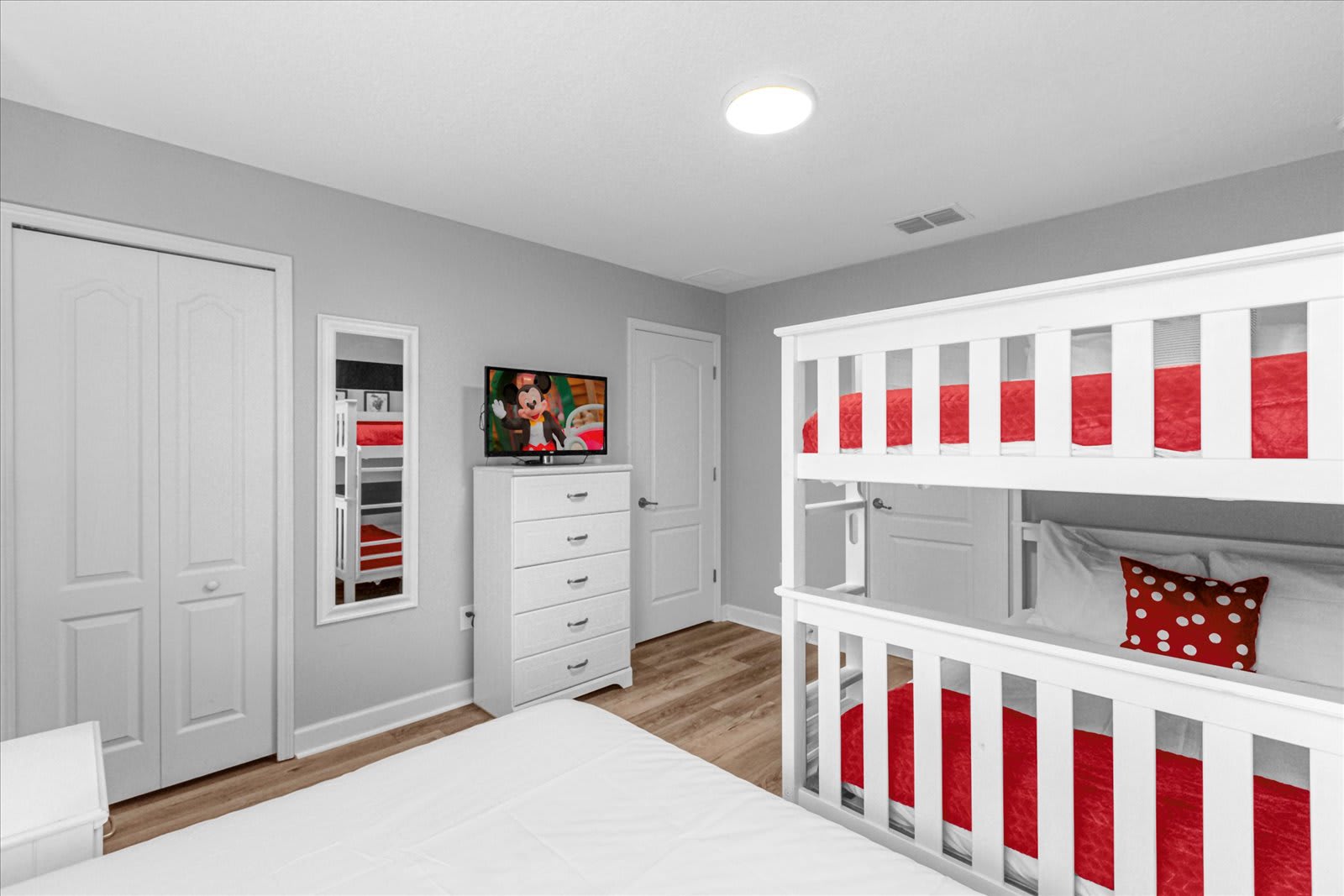 Mickey Mouse themed bedroom showing a white dresser with a TV, a full-length mirror, and a white bunk bed with red bedding, all on light wood-look flooring.