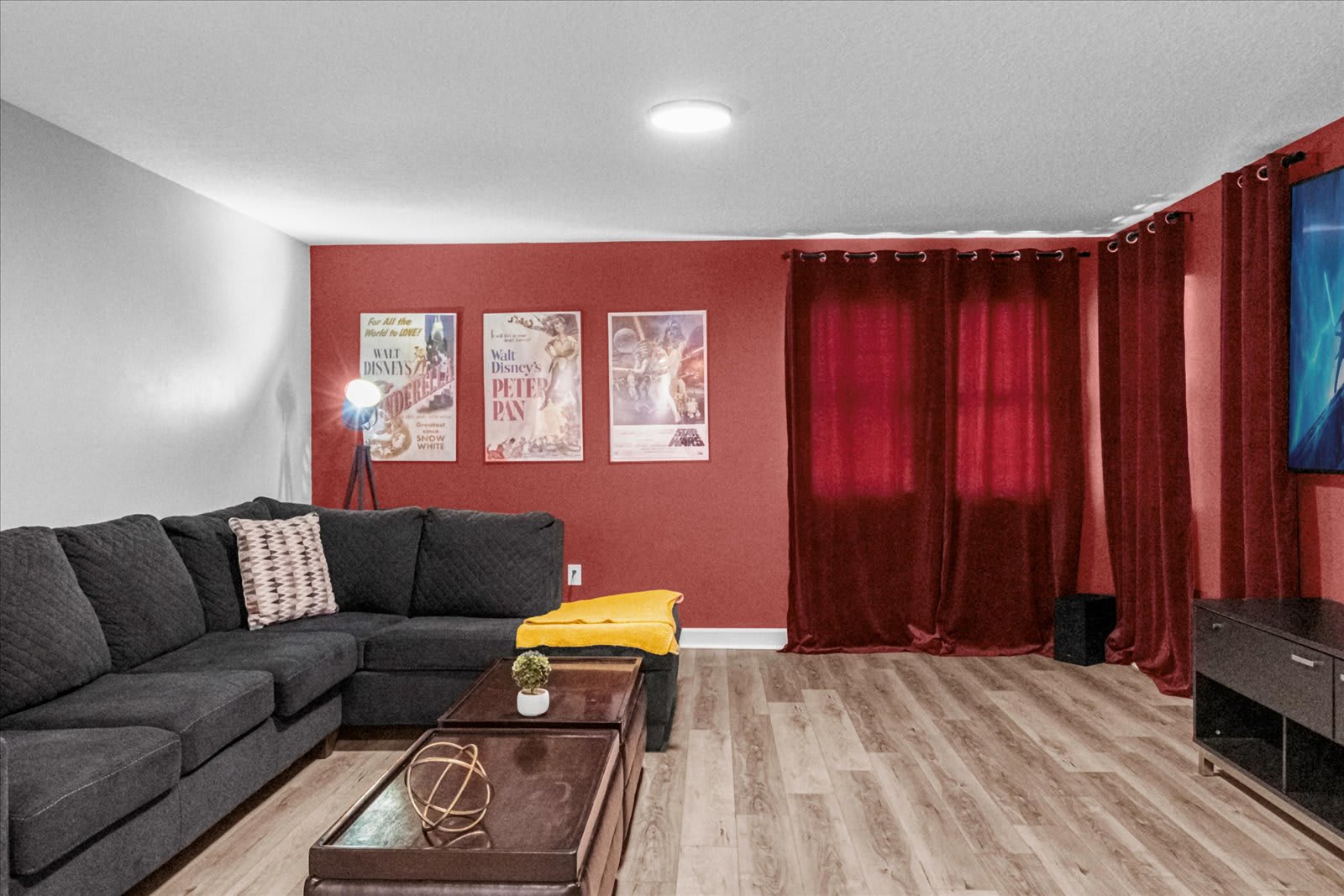 Another perspective of the media room, highlighting the dark sectional sofa with throw pillows and the two dark wood coffee tables. The red walls are decorated with vintage movie posters, and the room has wood laminate flooring.
