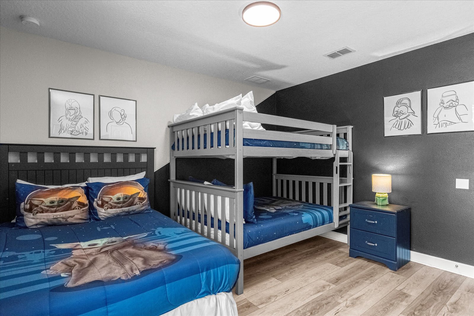 Star Wars themed bedroom with a queen bed featuring Grogu pillows and blue bedding, a grey bunk bed with blue bedding, and black and white character art on the dark accent wall.