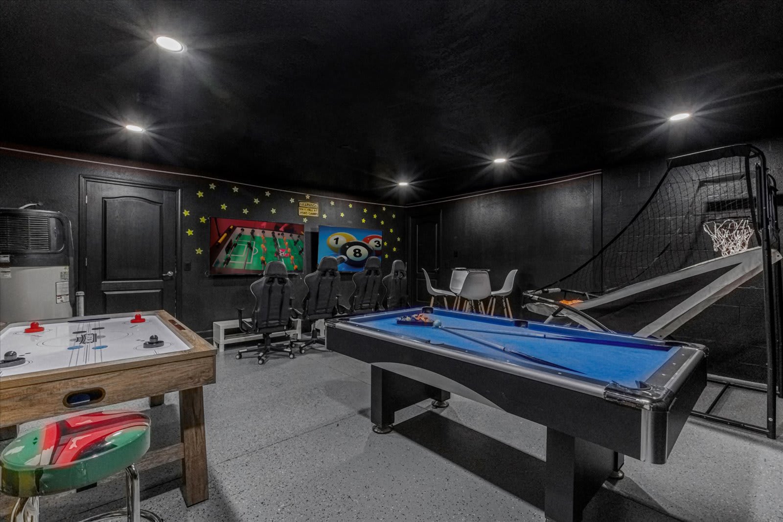 A vibrant game room with dark walls and ceiling, featuring an air hockey table, a blue-felt pool table, and a basketball arcade game. A gaming setup with multiple screens and chairs is against a wall decorated with star decals.
