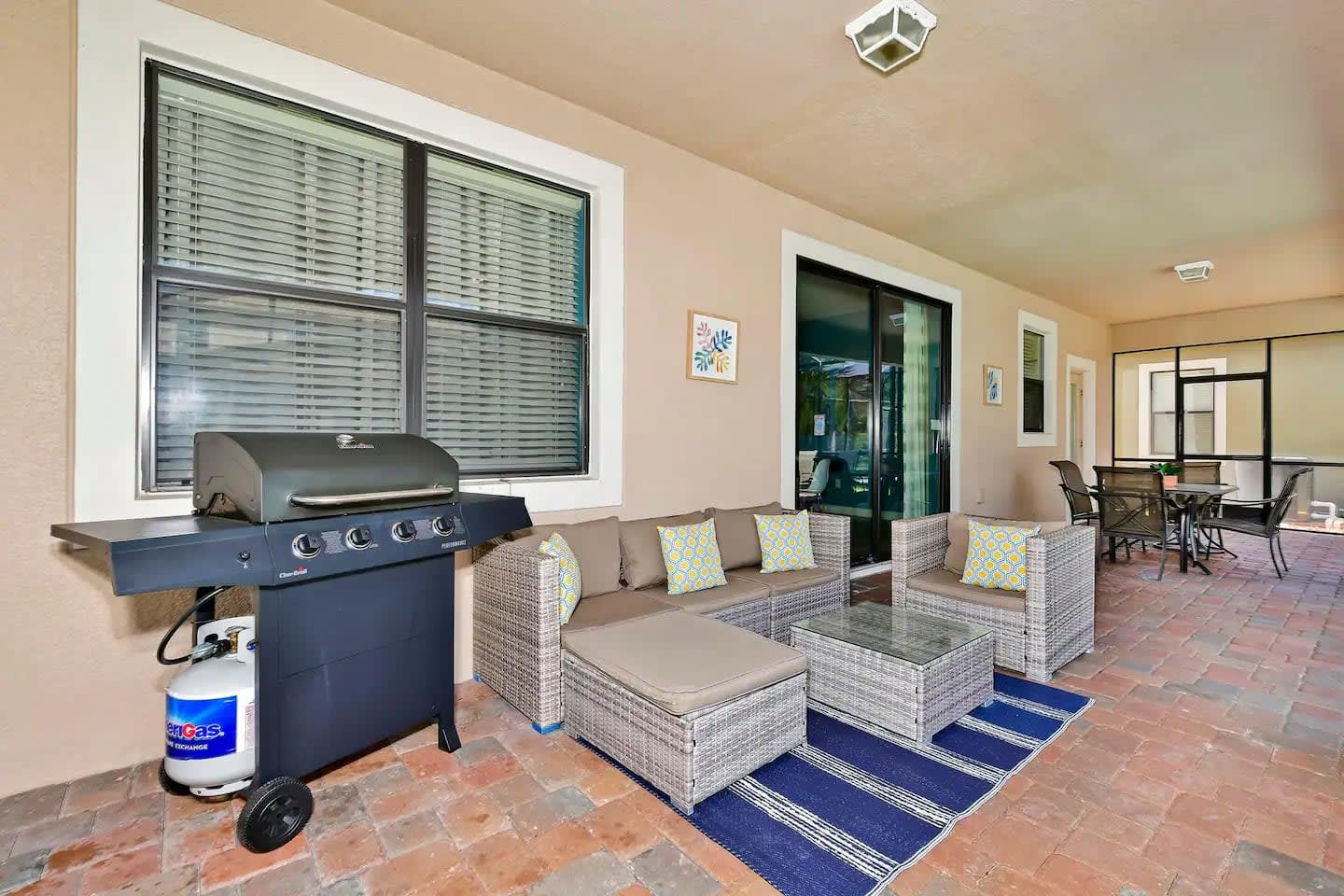 A covered outdoor patio area with brick pavers, featuring a wicker sectional sofa with cushions and a glass-top coffee table. A black BBQ grill with a propane tank is positioned against the house wall, with large windows and sliding glass doors leading indoors.
