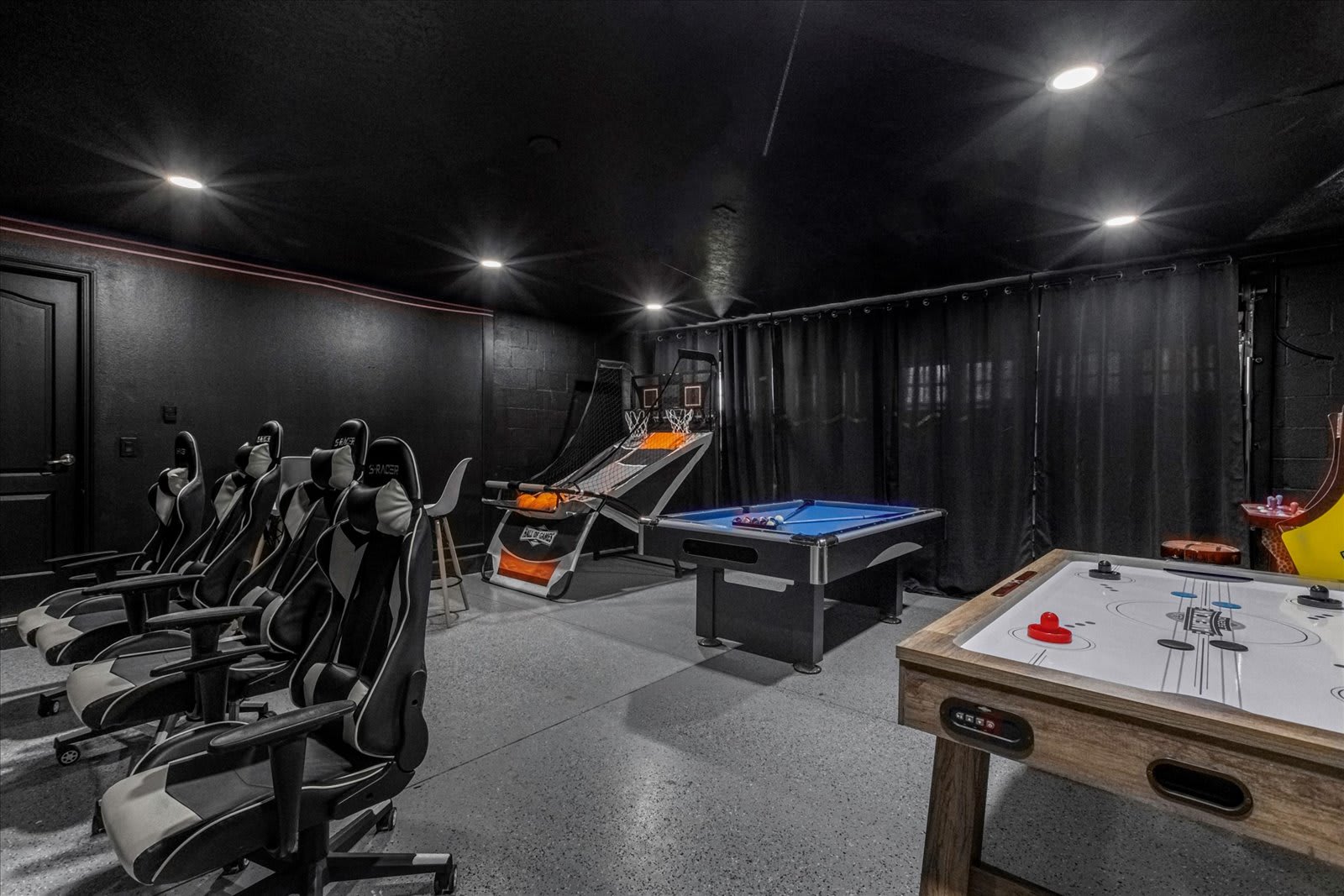 Dark-themed game room with black walls and ceiling, featuring a row of gaming chairs, a basketball arcade game, a pool table with blue felt, and an air hockey table. Recessed lighting illuminates the various game stations.